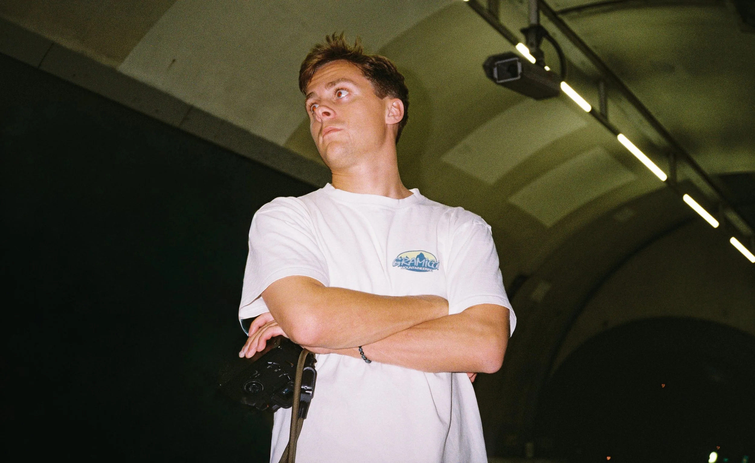 Ung mann med kort hår, kledd i hvitt T-skjorte med logo, står med krysset armer i et tunnel. Han ser oppover med en alvorlig utseende.