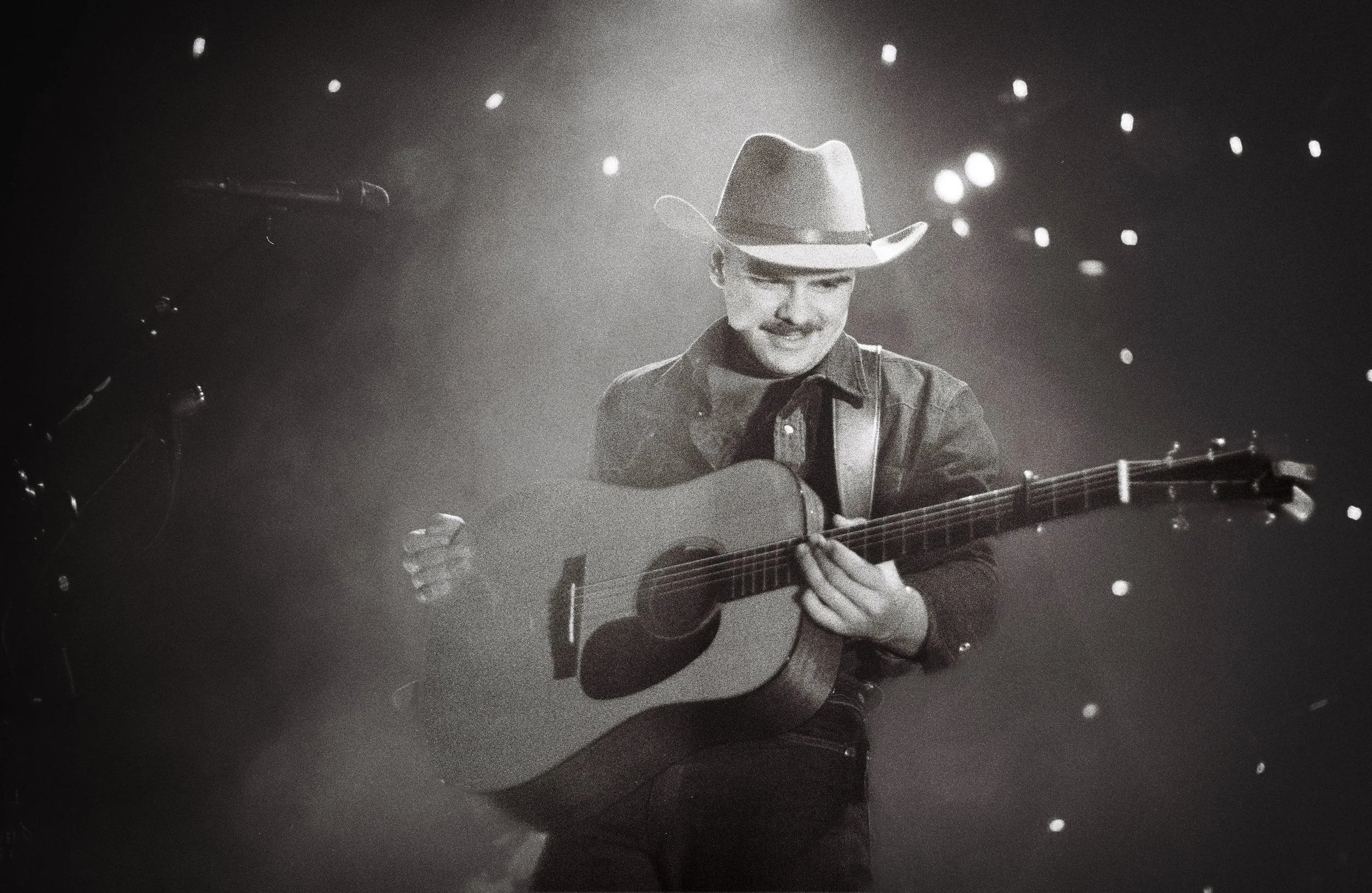 Black and white photo of a man wearing a cowboy hat and denim jacket playing an acoustic guitar on stage with lights in the background.