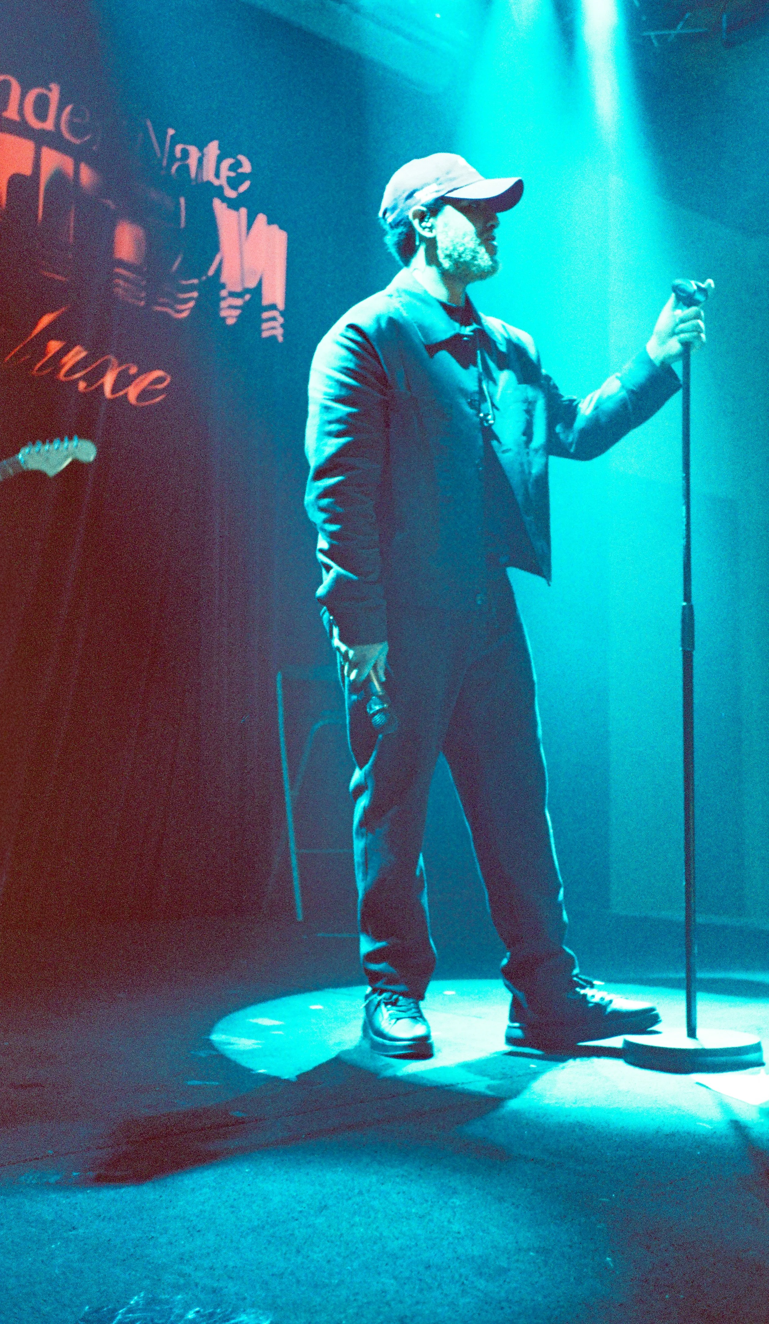 En mann på scenen med mikrofon i hånden, opplyst av blått lys, med en bakgrunn som har tekster og en gitar.