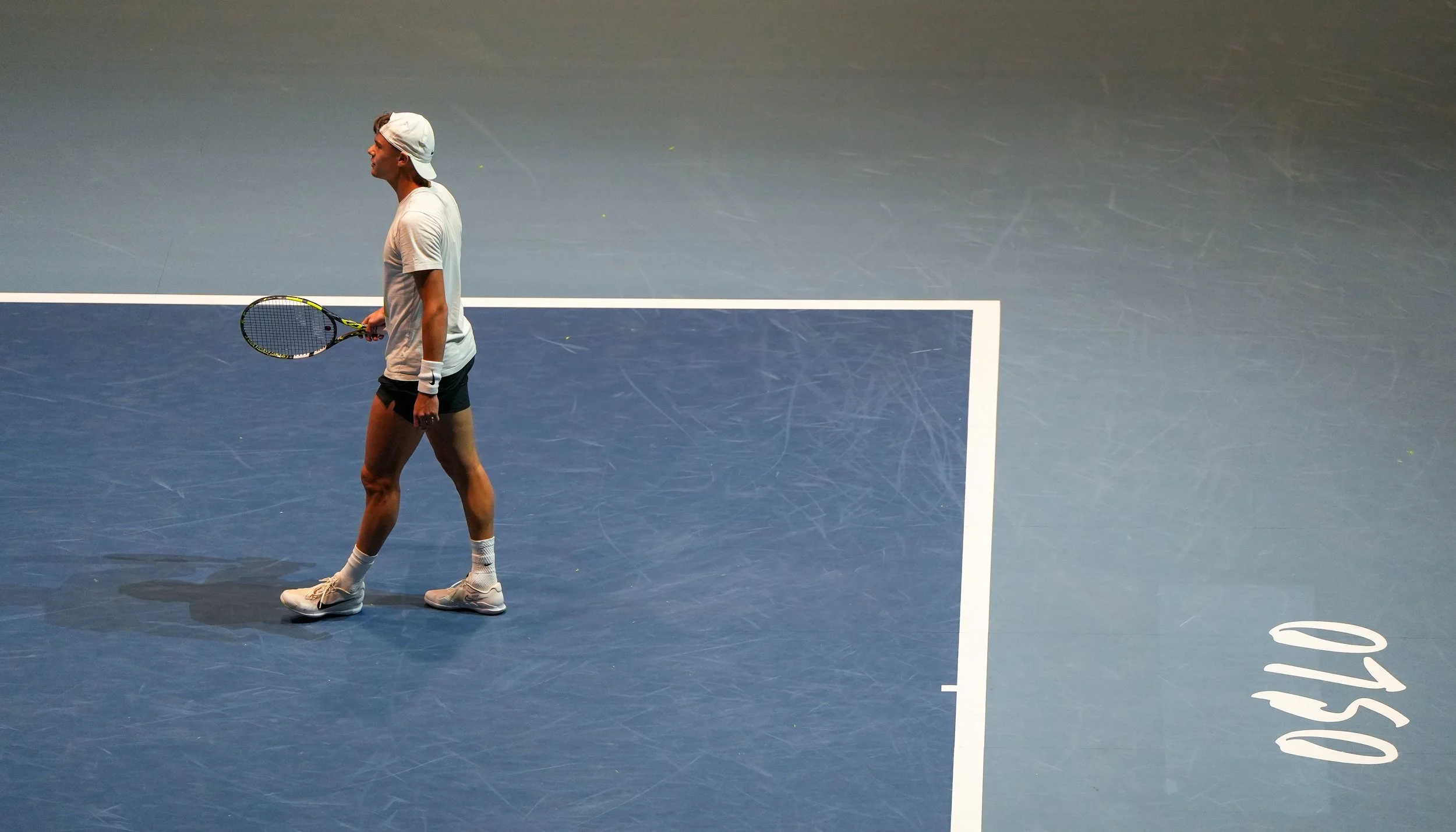 En mann på tennisbane, klar for spill, med tennisracket i hånden, iført hvit t-skjorte, svart shorts, og hvite sneakers, med en hvit baseball-cap på hodet.