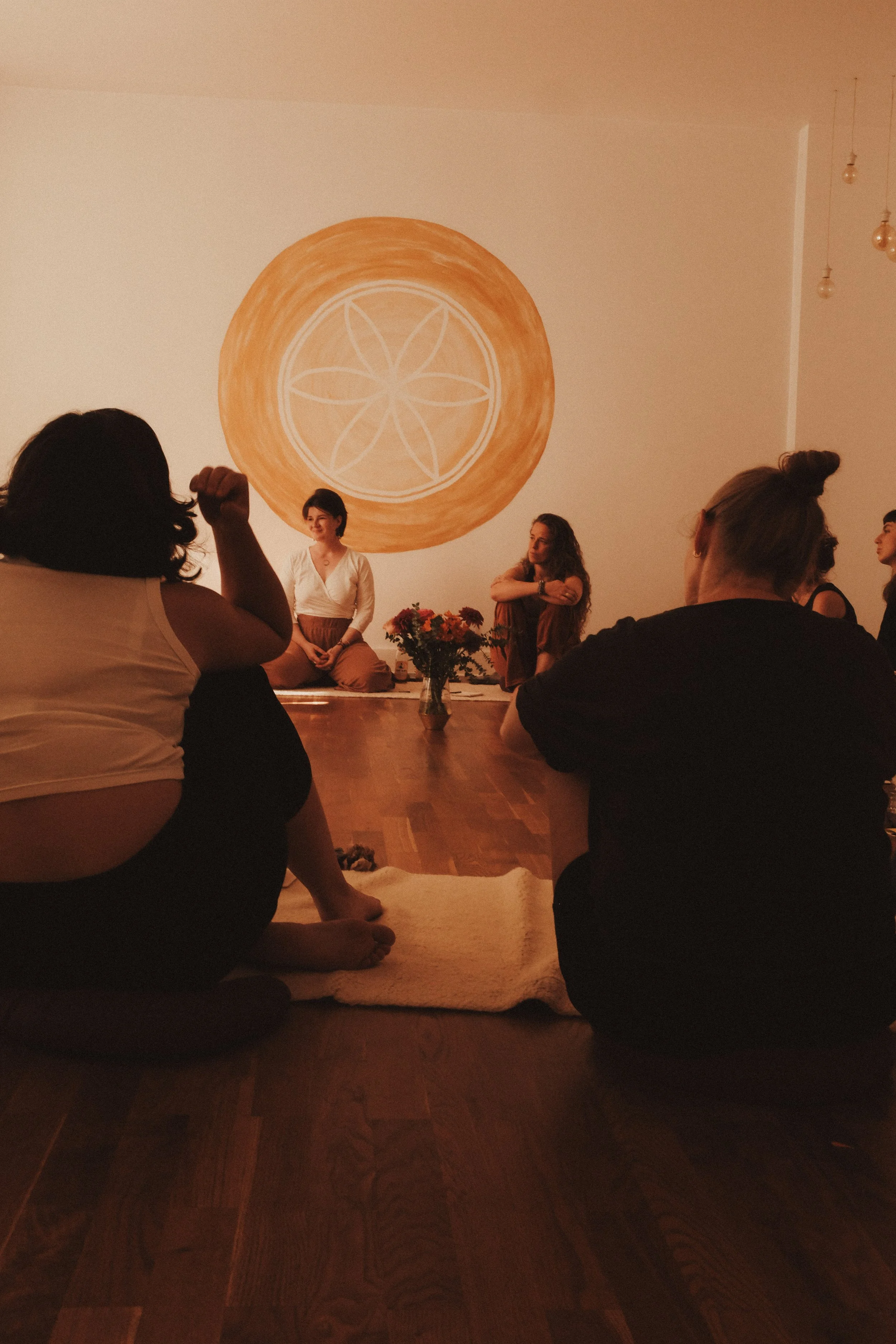 Eine Gruppe von Menschen sitzt im Halbdunkel auf dem Boden in einem Raum, während eine Frau auf einer Plattform sitzt. Die Wand im Hintergrund hat ein gelb-orangefarbenes Mandala-Design.