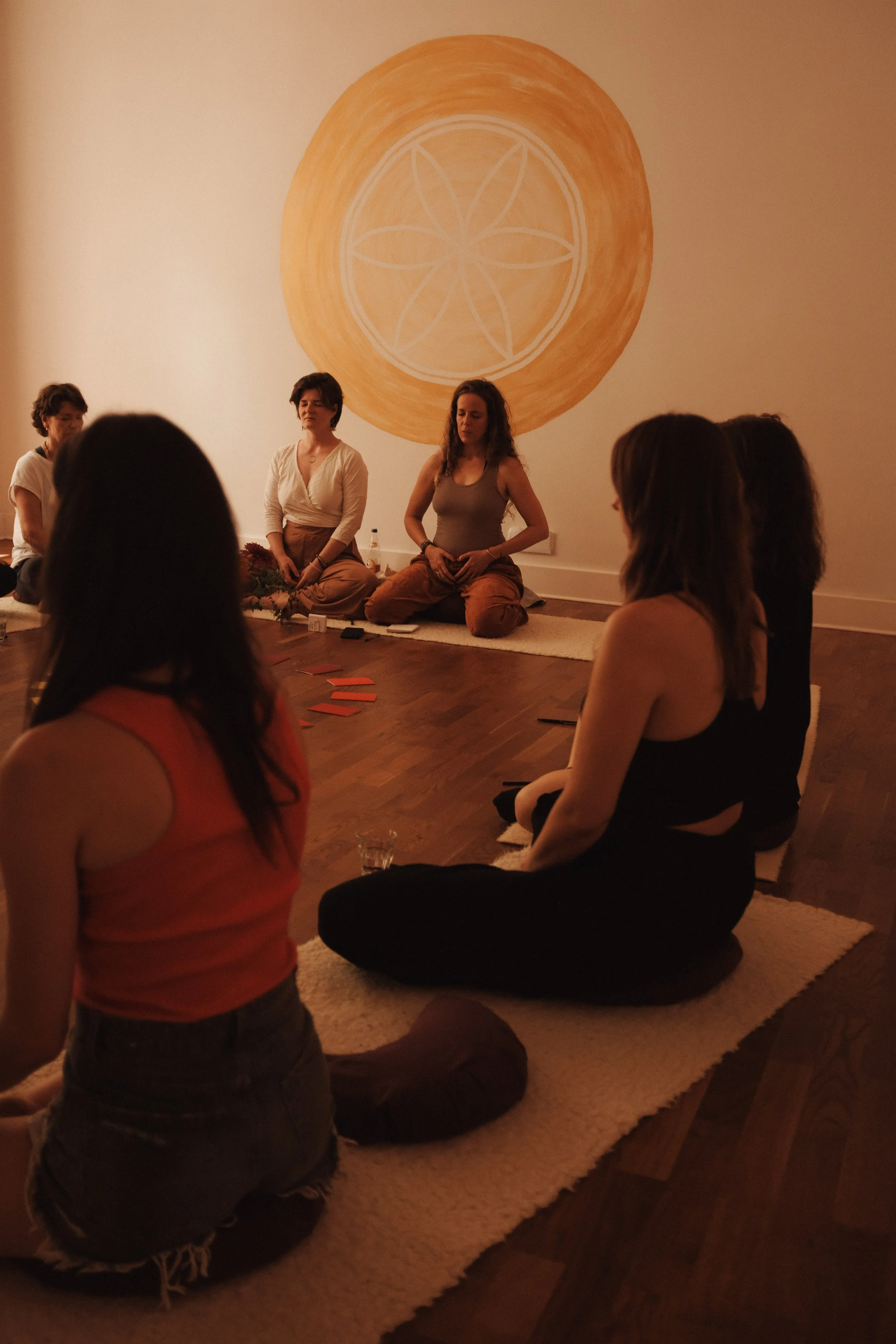 Menschen sitzen im Kreis auf Teppichen in einem Raum, meditieren oder nehmen an einer Sitzung teil, im Hintergrund ist eine Wand mit einem großen, orangefarbenen, runden Mandala oder Symbol.