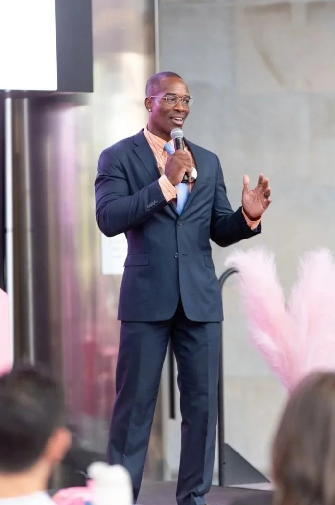 A man in a dark suit, pink shirt, and glasses is speaking at a conference or event, holding a microphone in his right hand and gesturing with his left hand. There are blurred audience members and pink decorative feathers in the background.