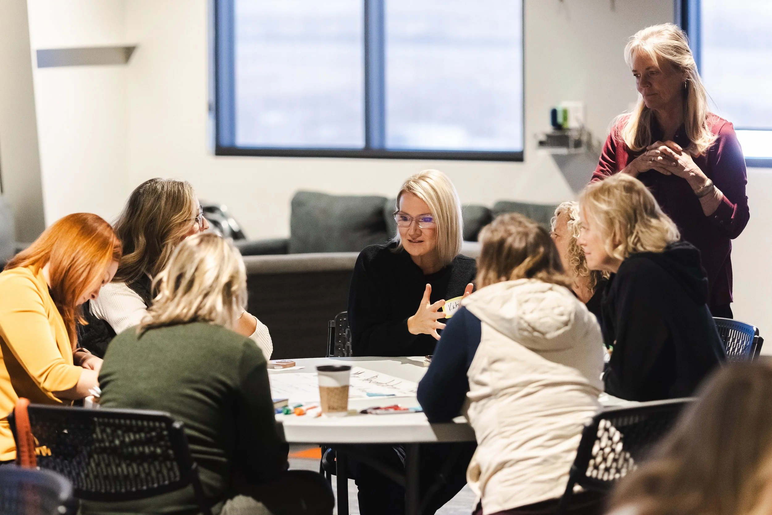 A group of women seated around a table in a meeting room, engaged in conversation, with a woman standing nearby. The atmosphere appears collaborative and professional.
