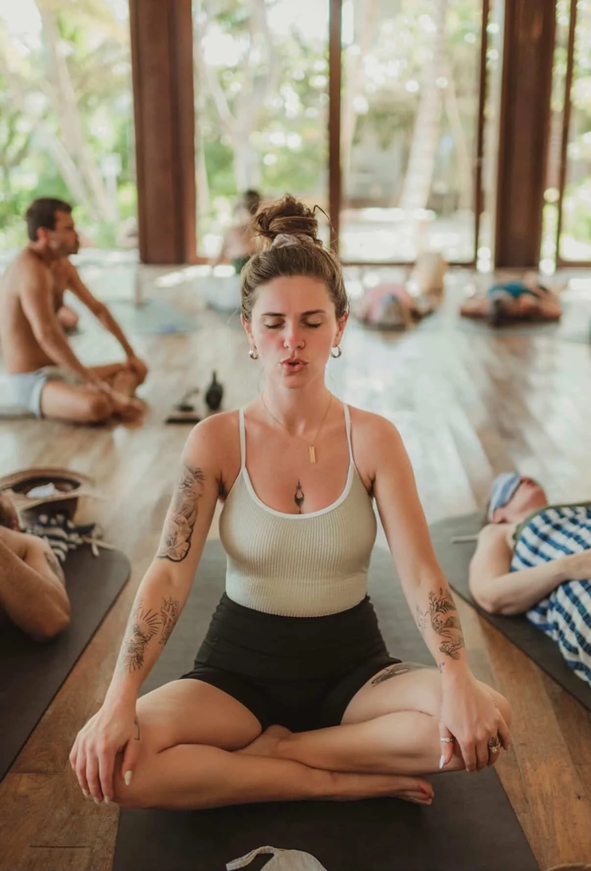 A woman with tattoos doing yoga in a seated meditation pose in a spacious, well-lit room with large windows and natural scenery outside.