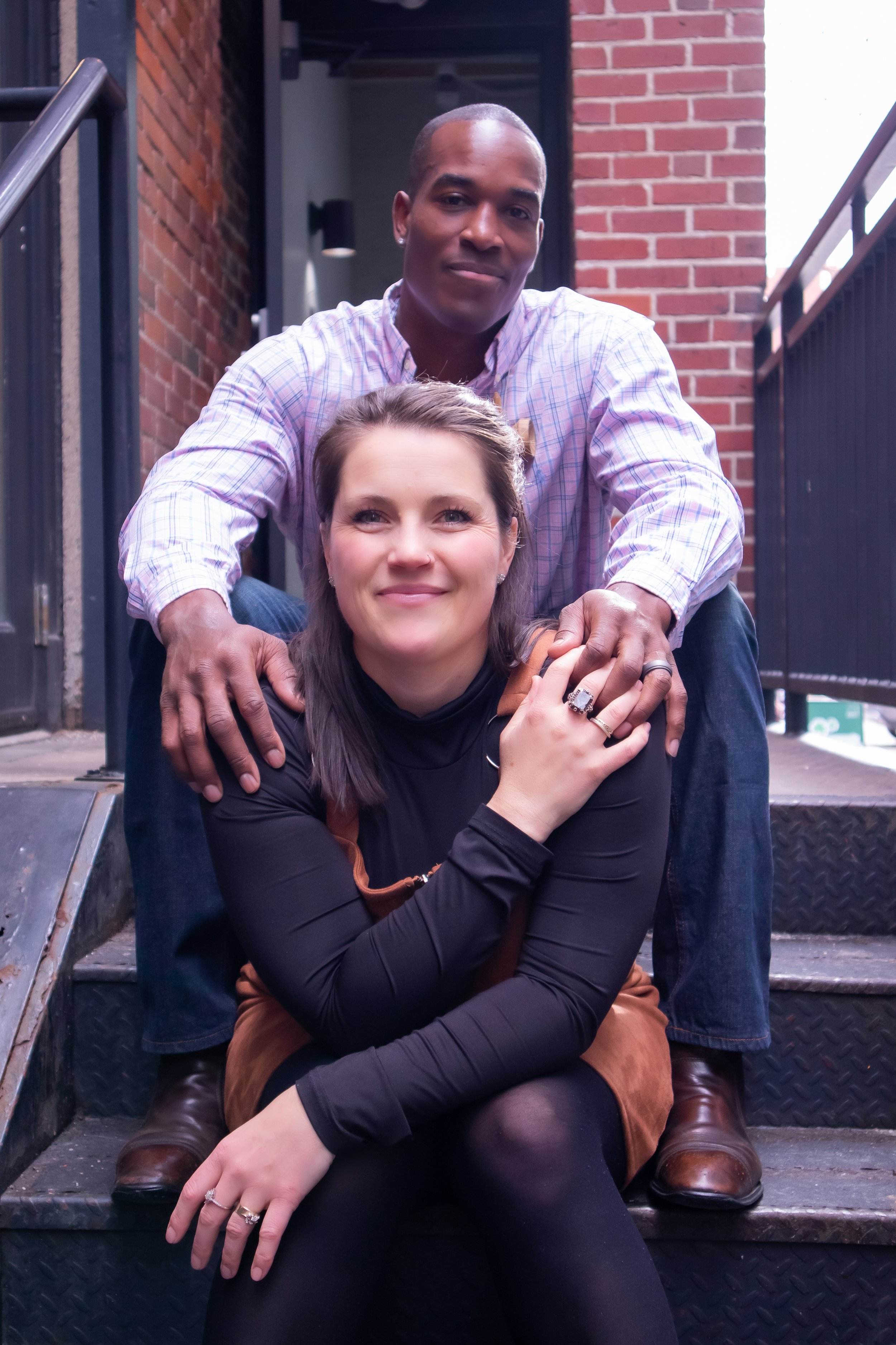 Two people sitting on outdoor steps in an urban setting, with brick walls and metal railings, smiling and holding hands.
