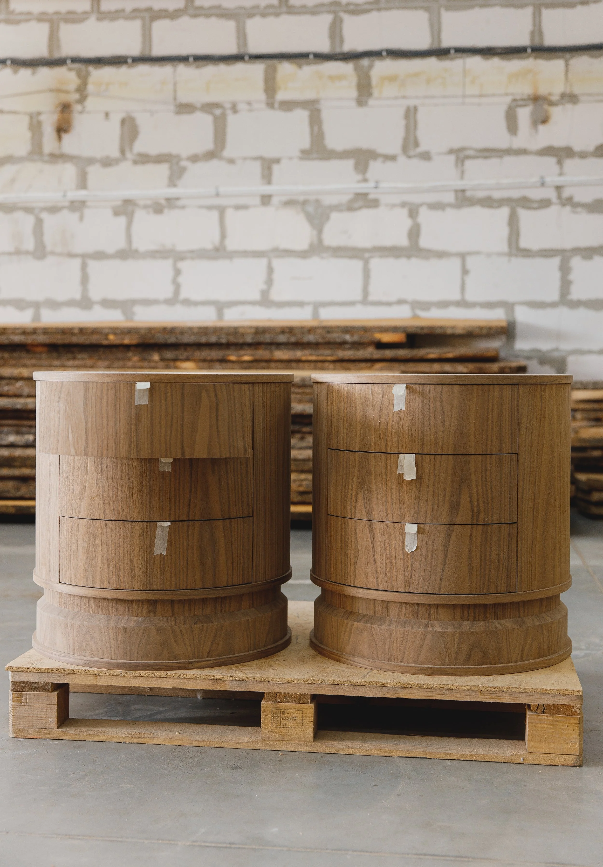 Two round wooden furniture pieces with three drawers each, placed on a wooden pallet in a workshop setting with a brick wall in the background.