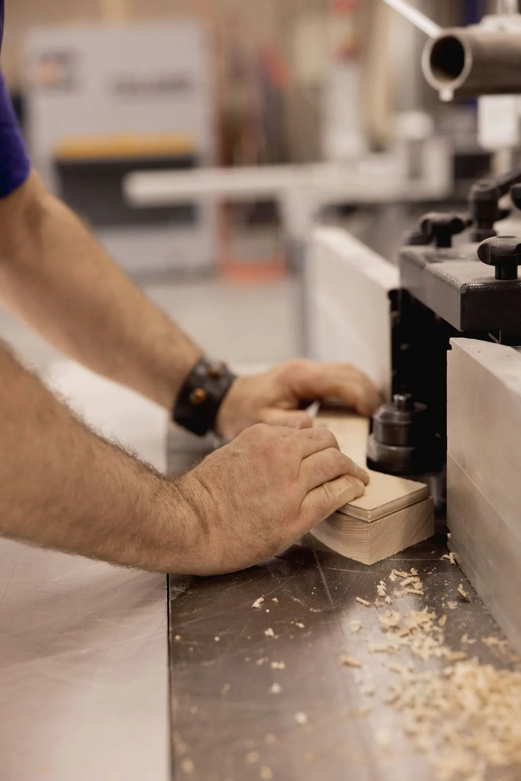 A craftsman guides a wooden board through a woodworking machine in a workshop, with wood shavings on the table.