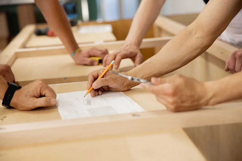 A team reviews a technical drawing on a wooden furniture frame, marking details with a pencil and measuring tools.