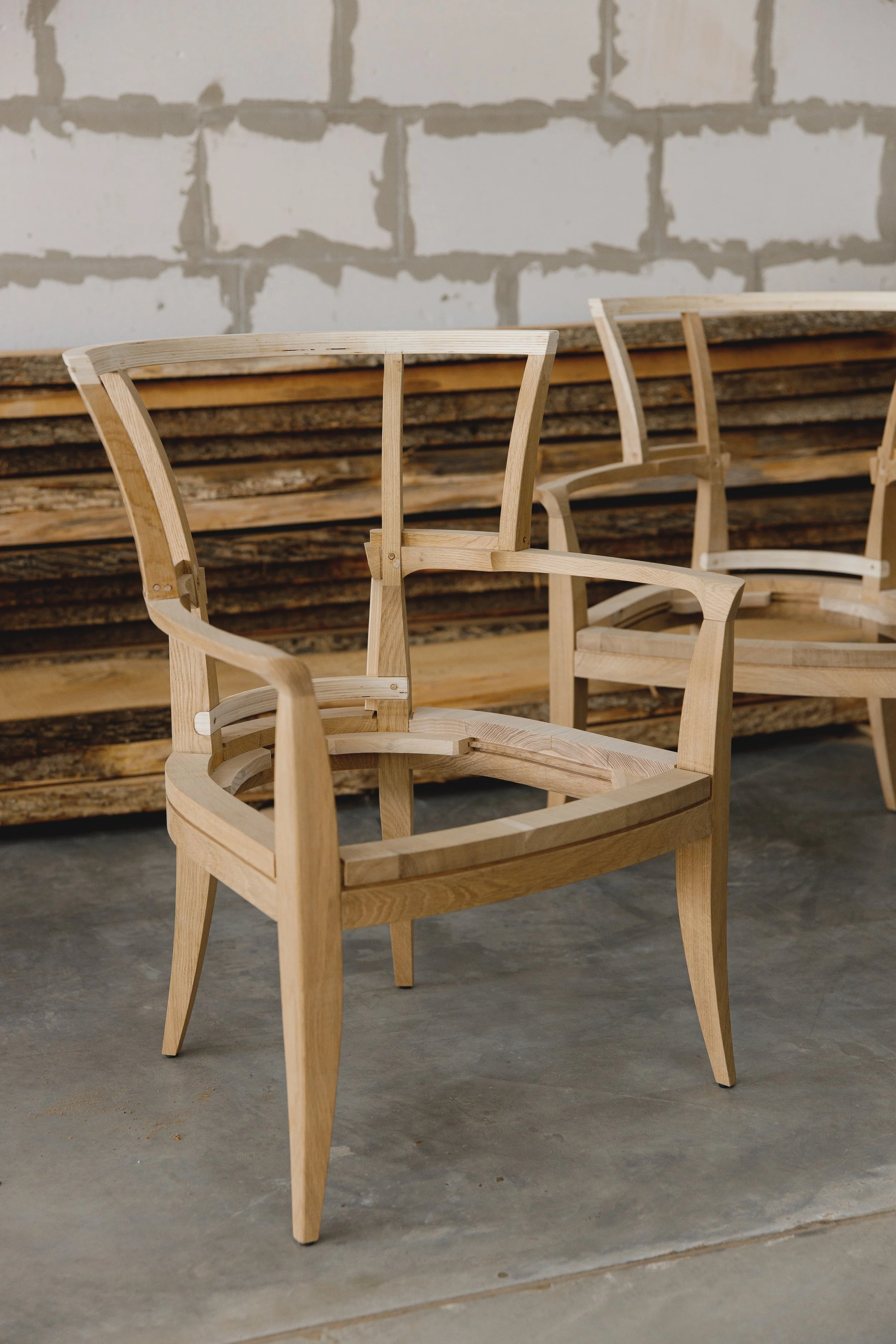Unfinished wooden armchair in a workshop with stacked wooden planks and a concrete floor.