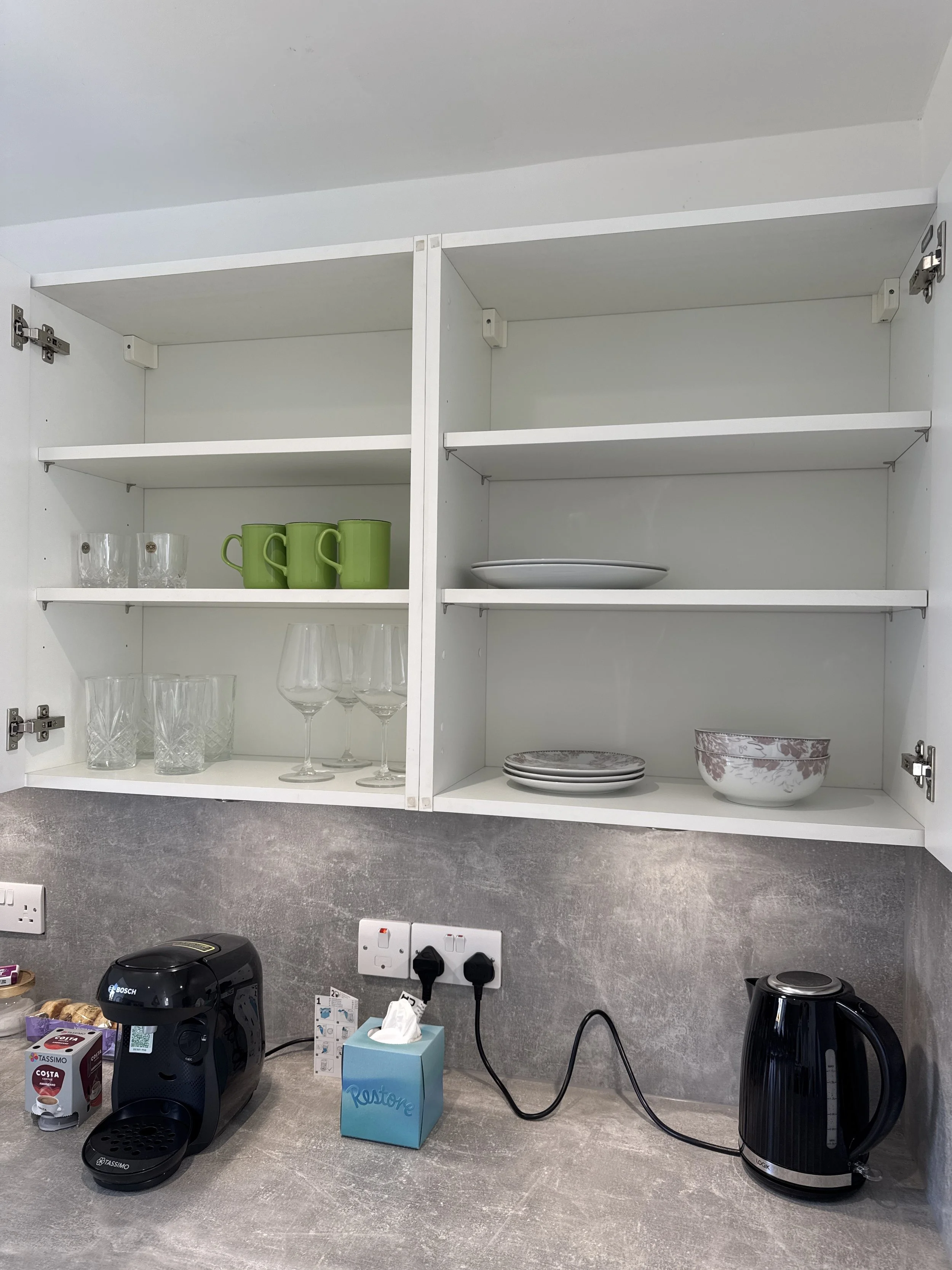Open white kitchen cabinet with glasses, green mugs, and bowls, a coffee machine, a tissue box, and an electric kettle on the countertop.