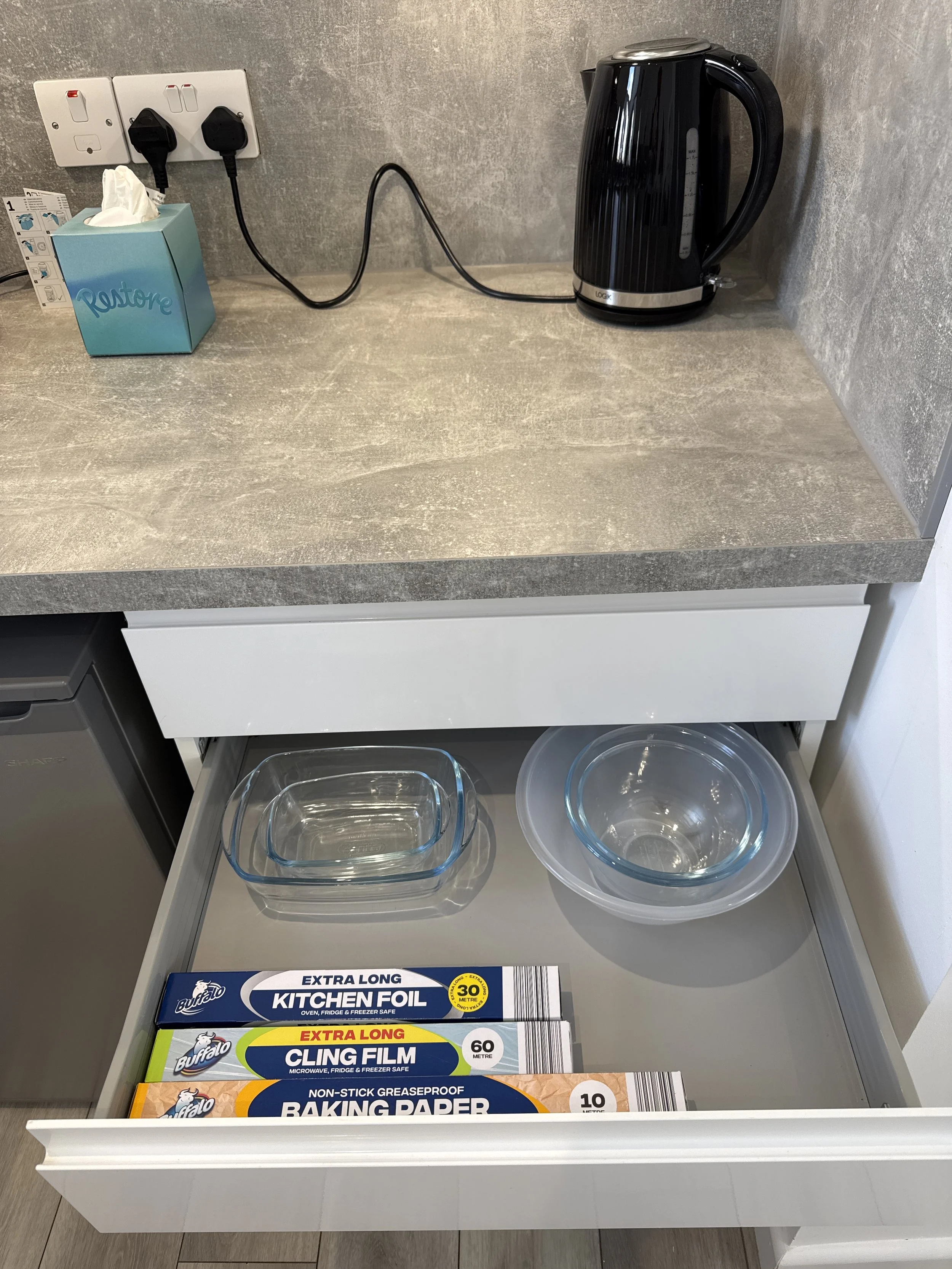 Open kitchen drawer containing glass baking dishes and foil, with a grey countertop, an electric kettle, tissue box, and electrical outlets on the wall above.