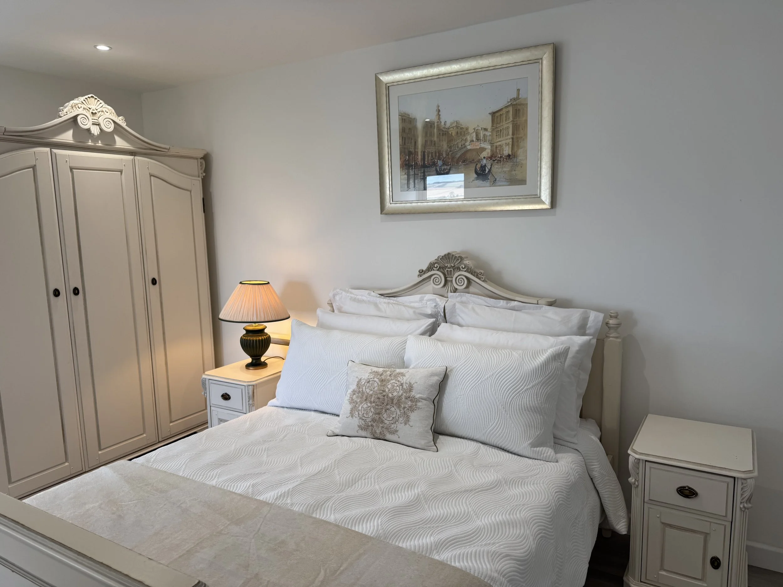 A cozy bedroom with a white ornate bed, matching nightstands, and a large wardrobe. The bed has white pillows and a decorative pillow with a floral pattern. A table lamp with a beige shade sits on one nightstand. Above the bed is a framed painting of