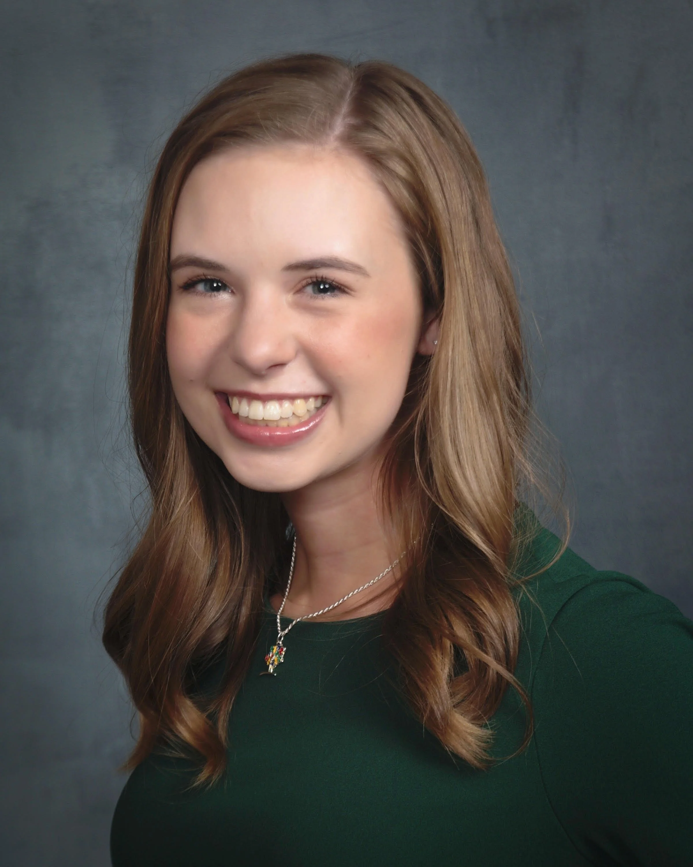 A young woman with long, wavy light brown hair smiling, wearing a dark green top, a silver necklace with a colorful pendant, and small earrings, against a dark gray background.