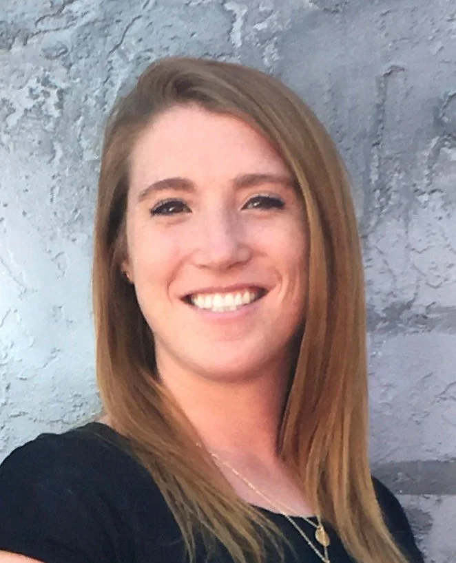 A woman with long light brown hair smiling at the camera, standing against a textured gray wall.