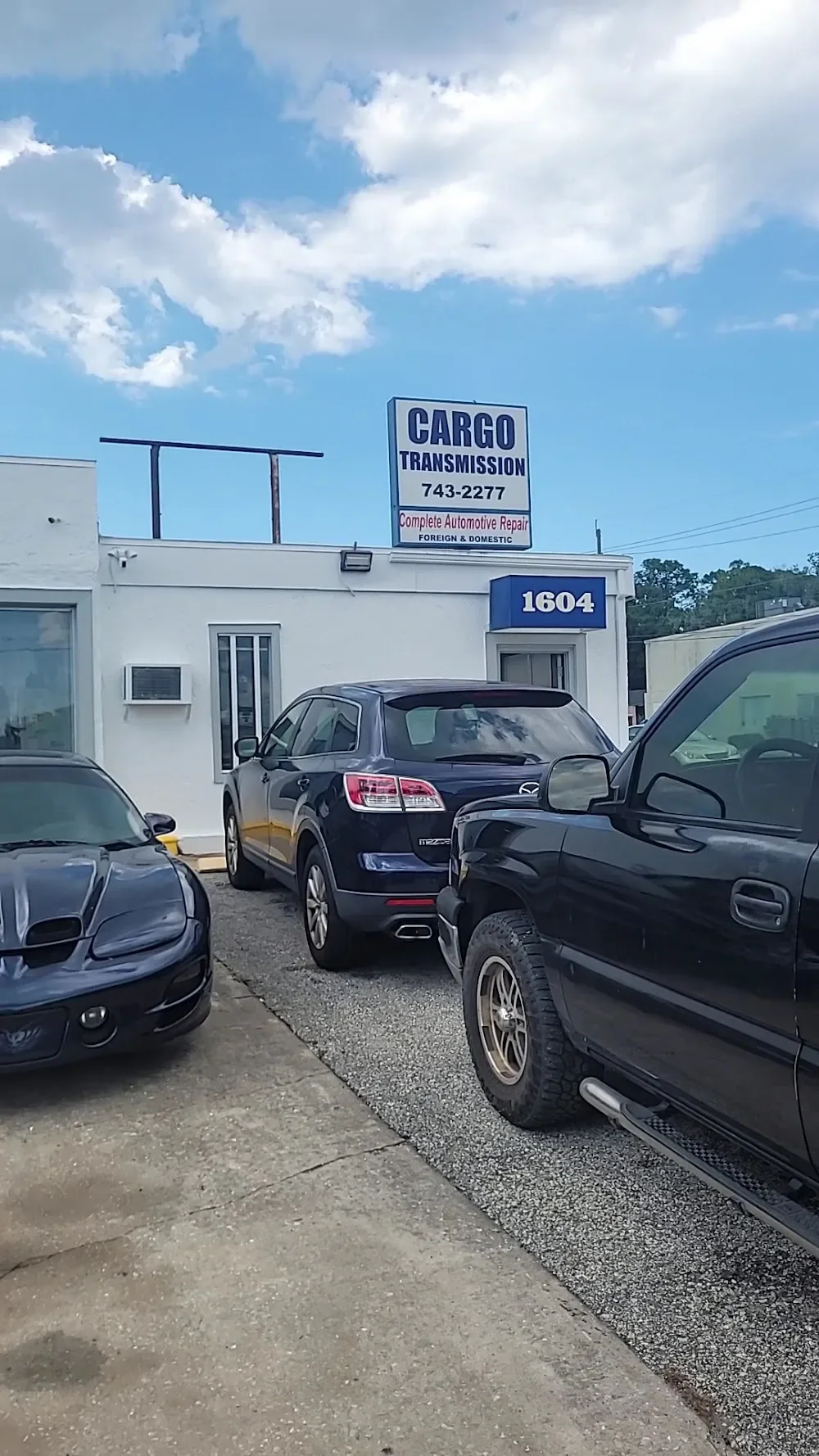 A small auto repair shop with a white exterior, marked with the address 1604. There are three parked cars in front of the shop, including a black Mazda SUV, a black Corvette sports car, and a black pickup truck. A sign above the building reads 'Cargo Transmission' and 'Complete Automotive Repair,' with a phone number and indications of services for foreign and domestic vehicles.