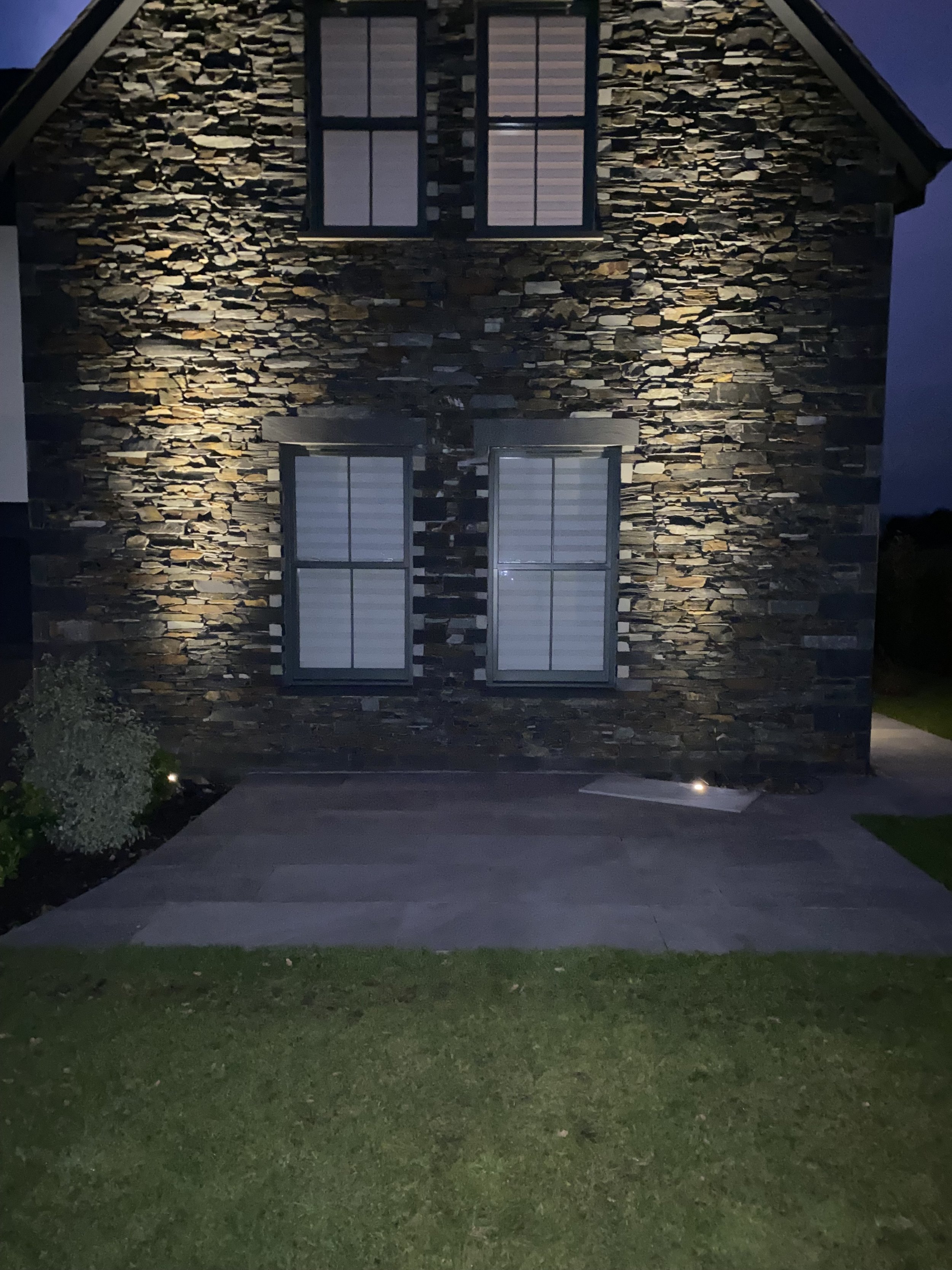 Nighttime view of a stone house with three windows illuminated from inside, outdoor lighting highlighting the stone facade, concrete pathway, and a small patch of grass in front.