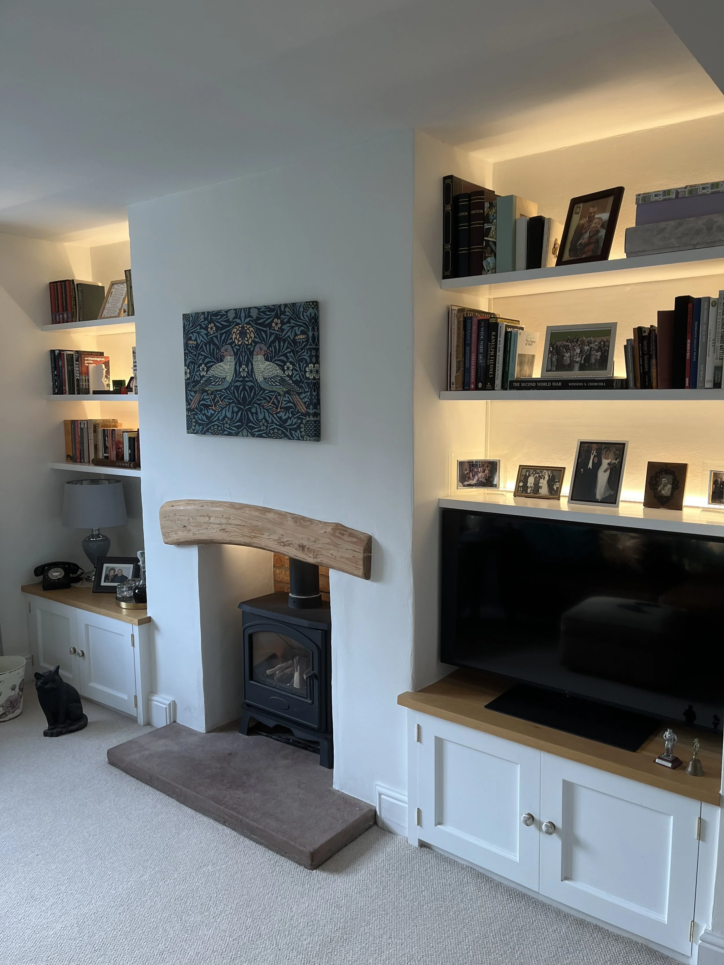 Living room corner with white built-in cabinets, a flat-screen TV, shelves with books and photos, a wood stove with a wooden mantle, a lamp, and a black cat sculpture on the floor.