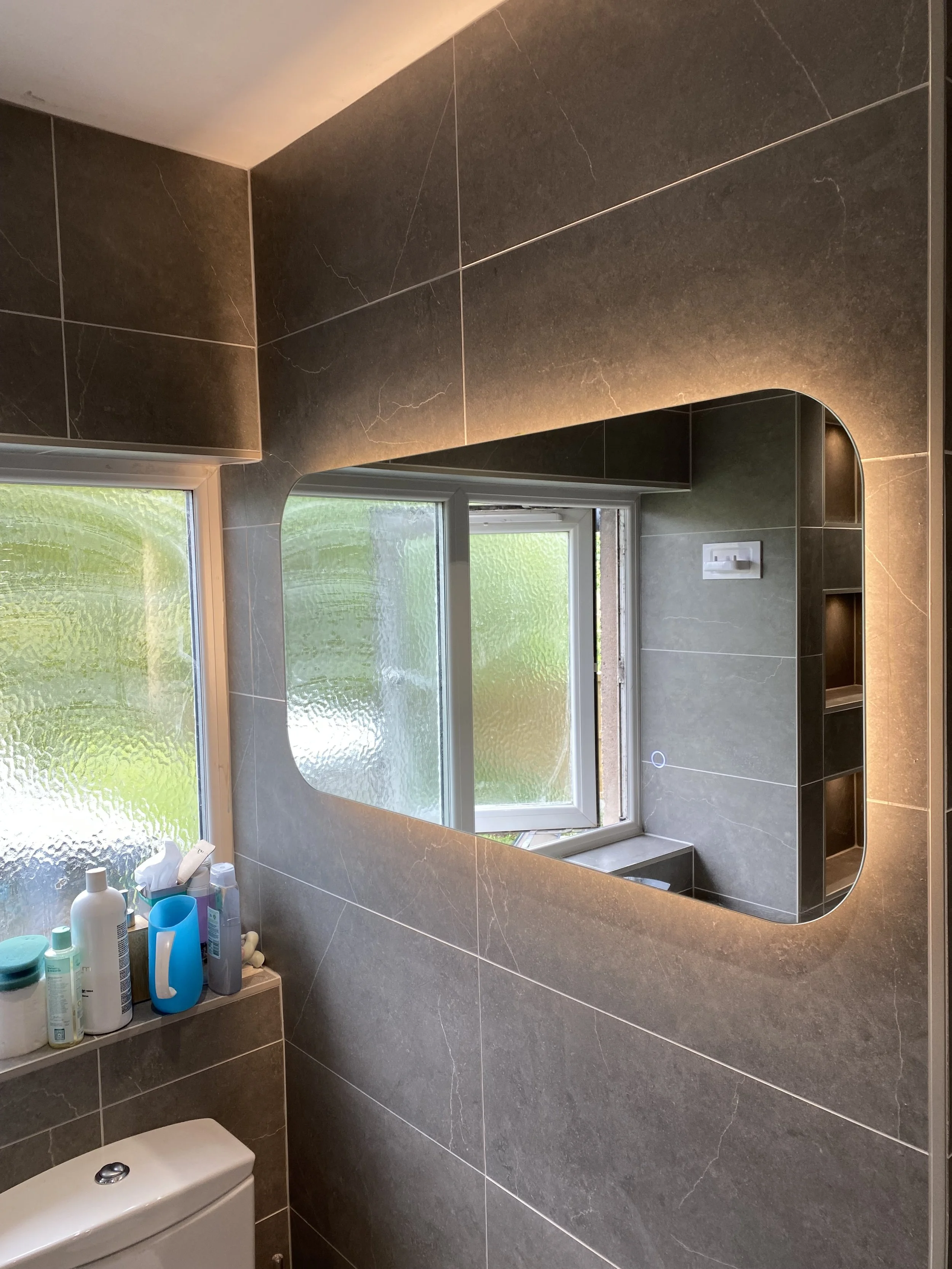 A bathroom with dark gray tiled walls, a frosted window, a large mirror with rounded corners, and a shelf with toiletries.