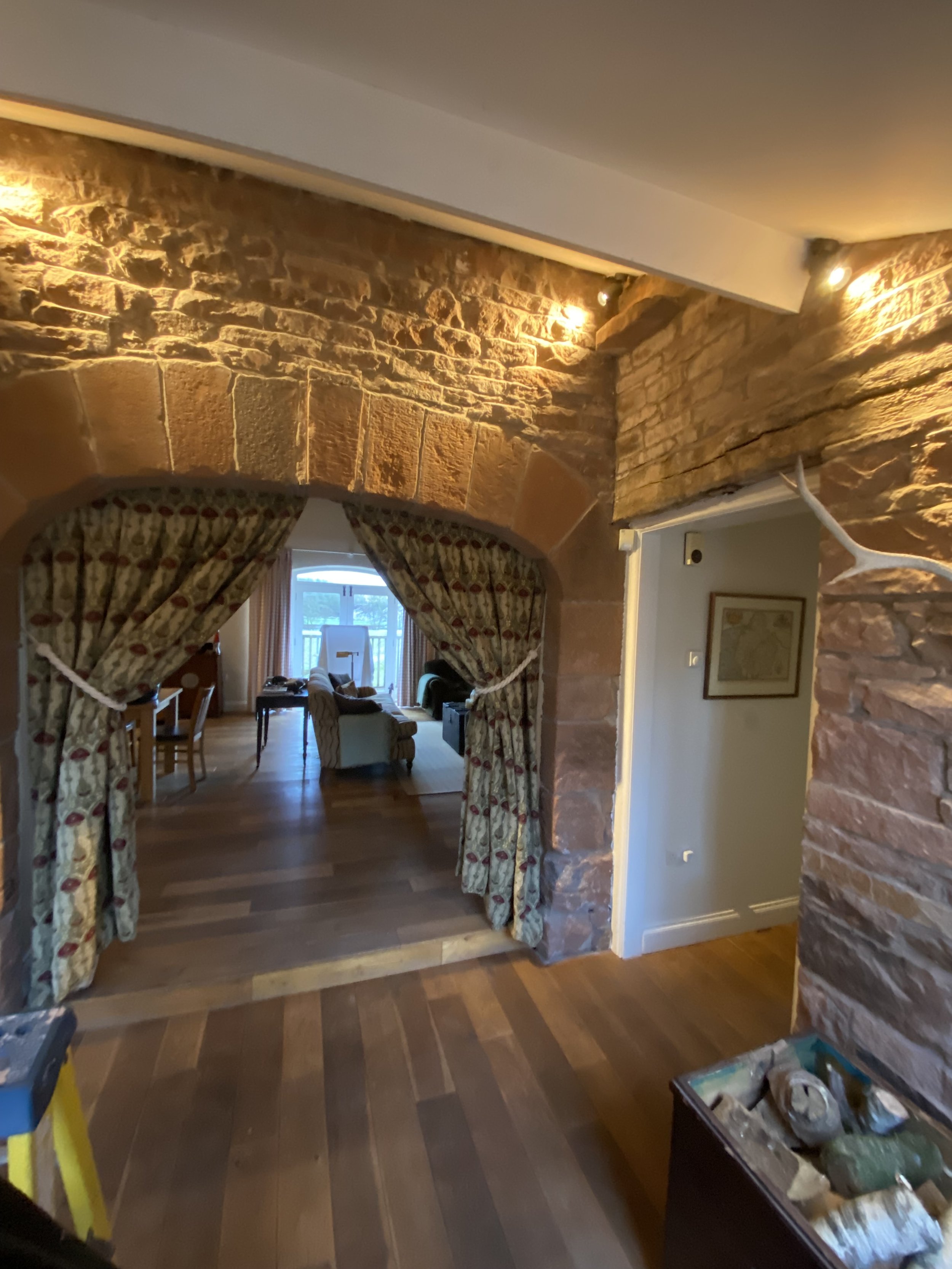 View of a living room with stone walls, wooden flooring, and large windows with floral curtains. The space is illuminated by warm lighting, and there are chairs, a sofa, and a table visible in the background.