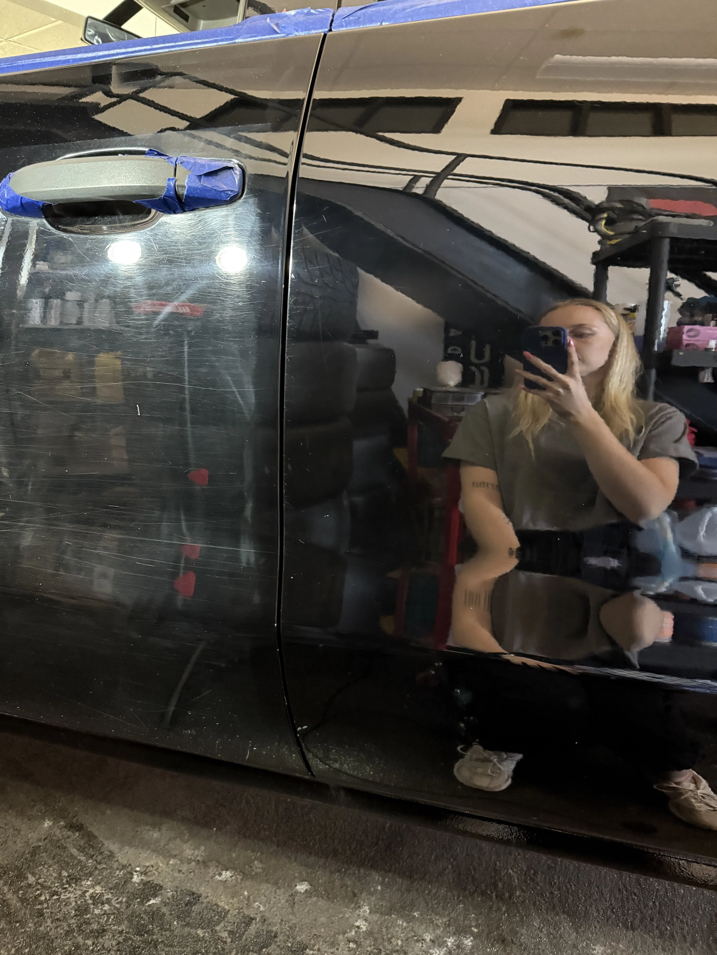 Reflection of a woman taking a photo with her phone in the shiny black side of a car, showing her blonde hair, gray T-shirt, and shorts, with a garage background including shelves, tools, and tires.
