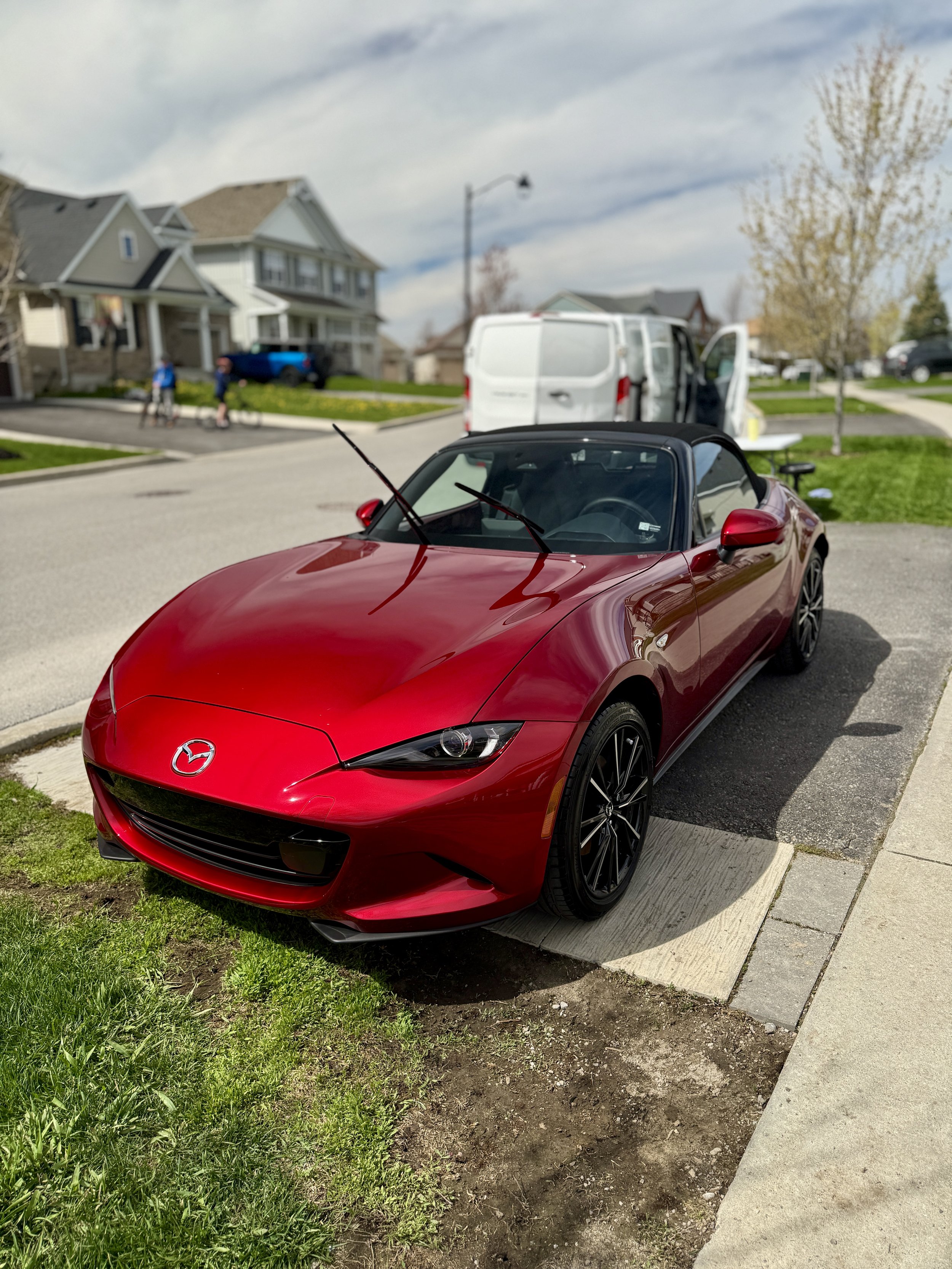 A red Mazda convertible parked on the street in front of a residential house. The house is a large, multi-story home with a porch and a driveway. There is a white van parked with its sliding door open and two people standing nearby. The neighborhood 
