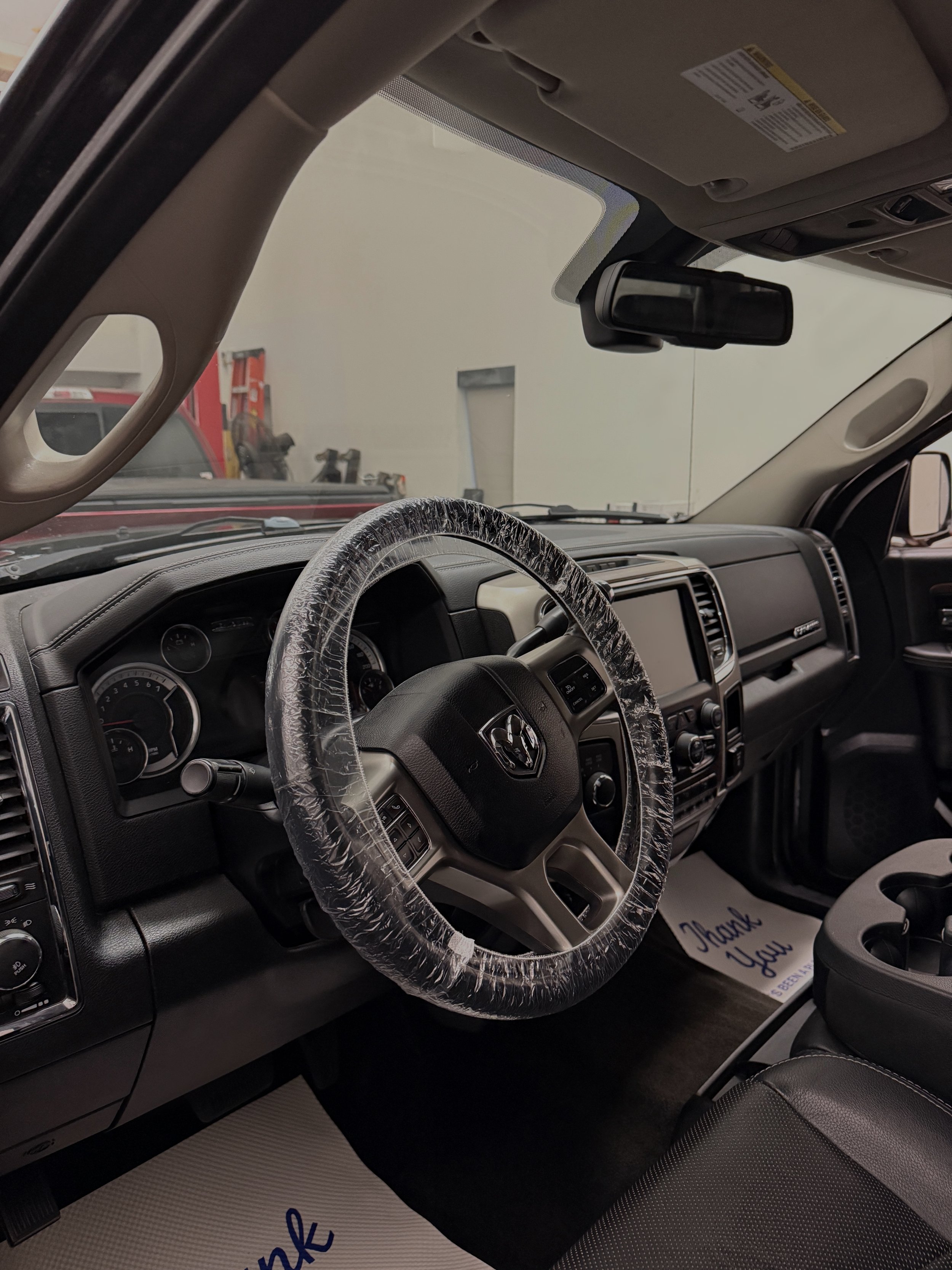 Interior of a vehicle, showing a plastic-wrapped steering wheel, dashboard, and driver-side seat with a paper mat that reads 'Thank You' on the floor.
