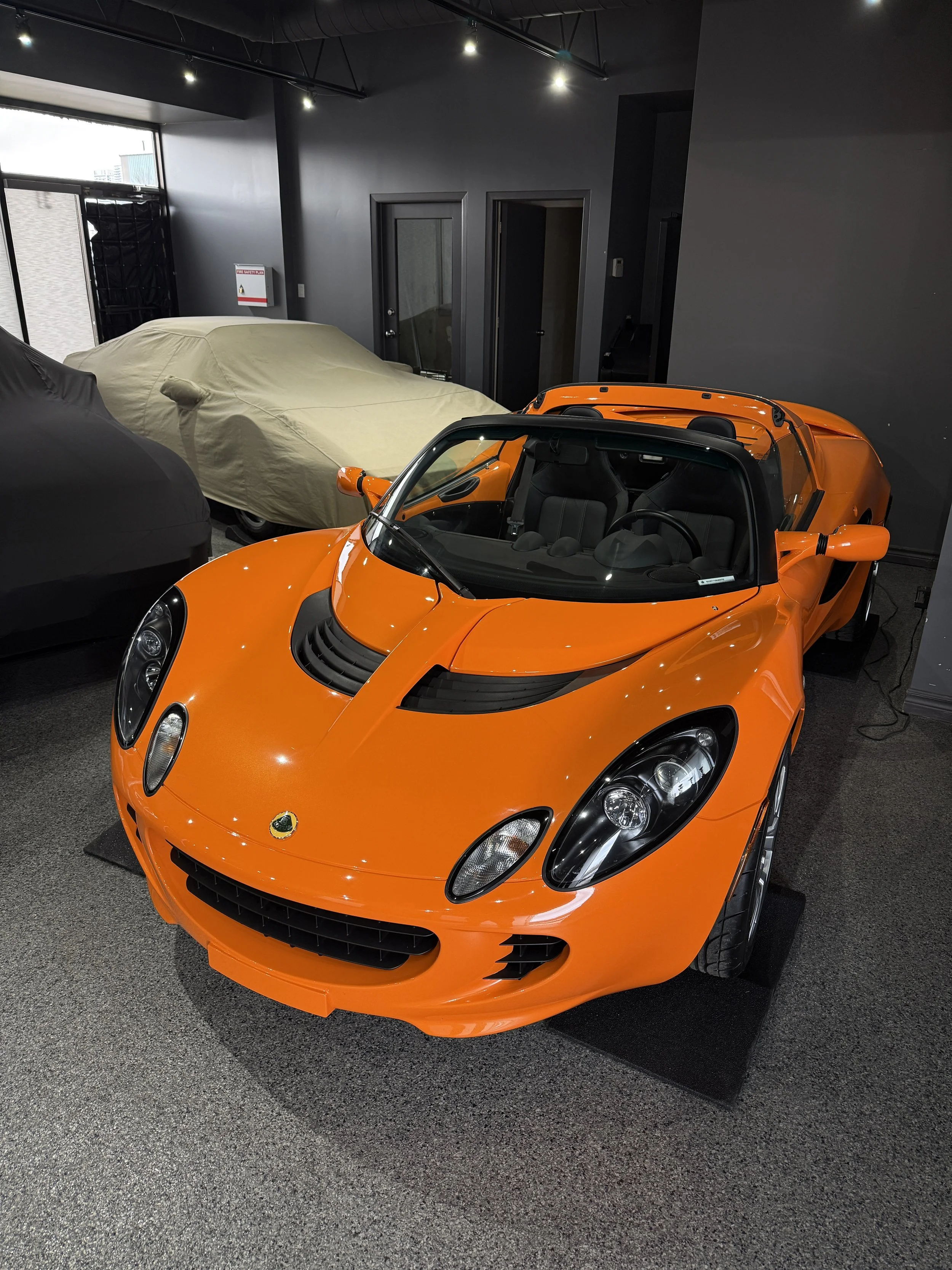 An orange sports car parked indoors, with a beige covered vehicle beside it and dark gray walls.