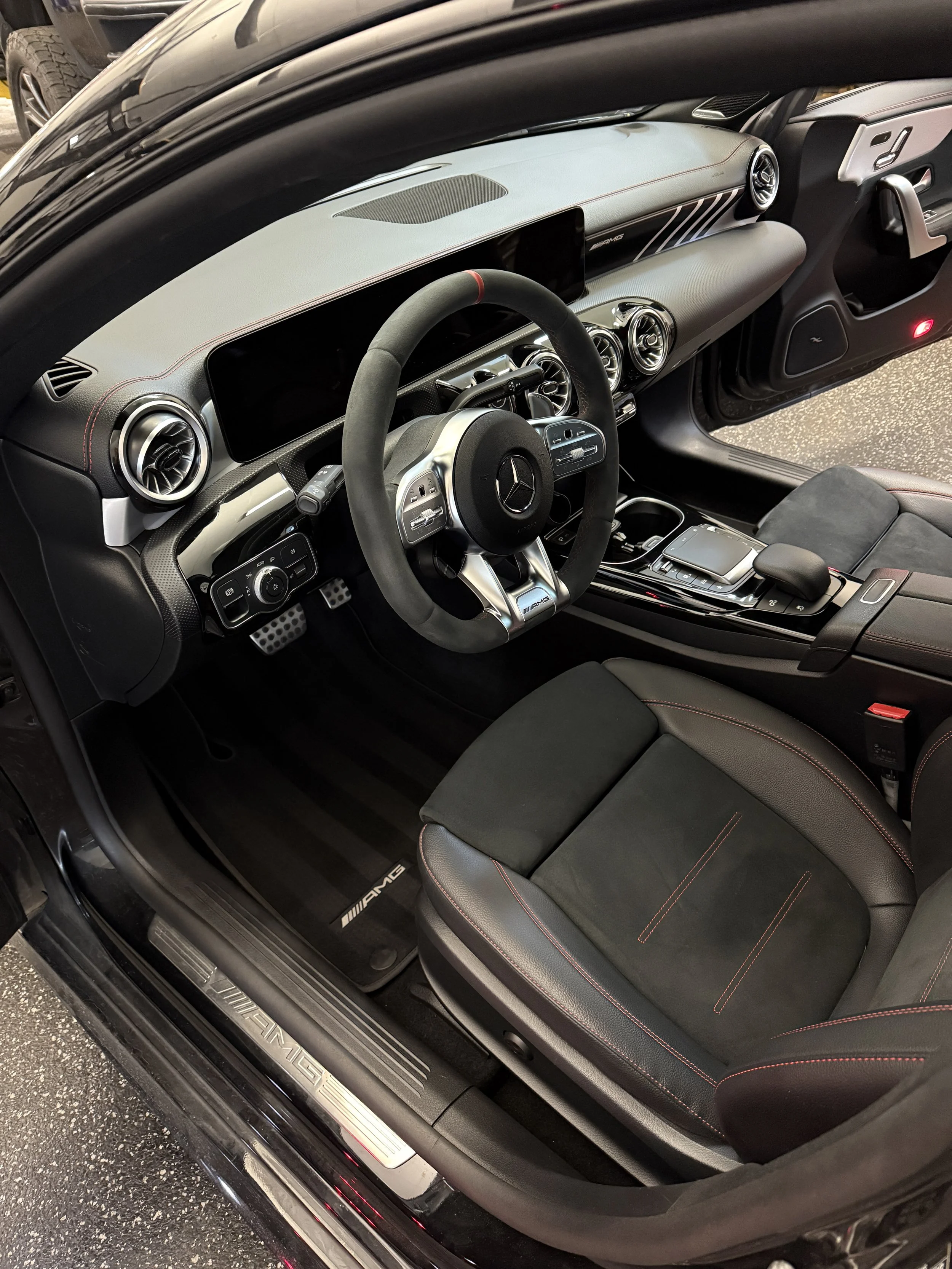 Interior view of a black Mercedes-Benz AMG car showing the dashboard, steering wheel, center console, and front seats with black upholstery and red stitching.