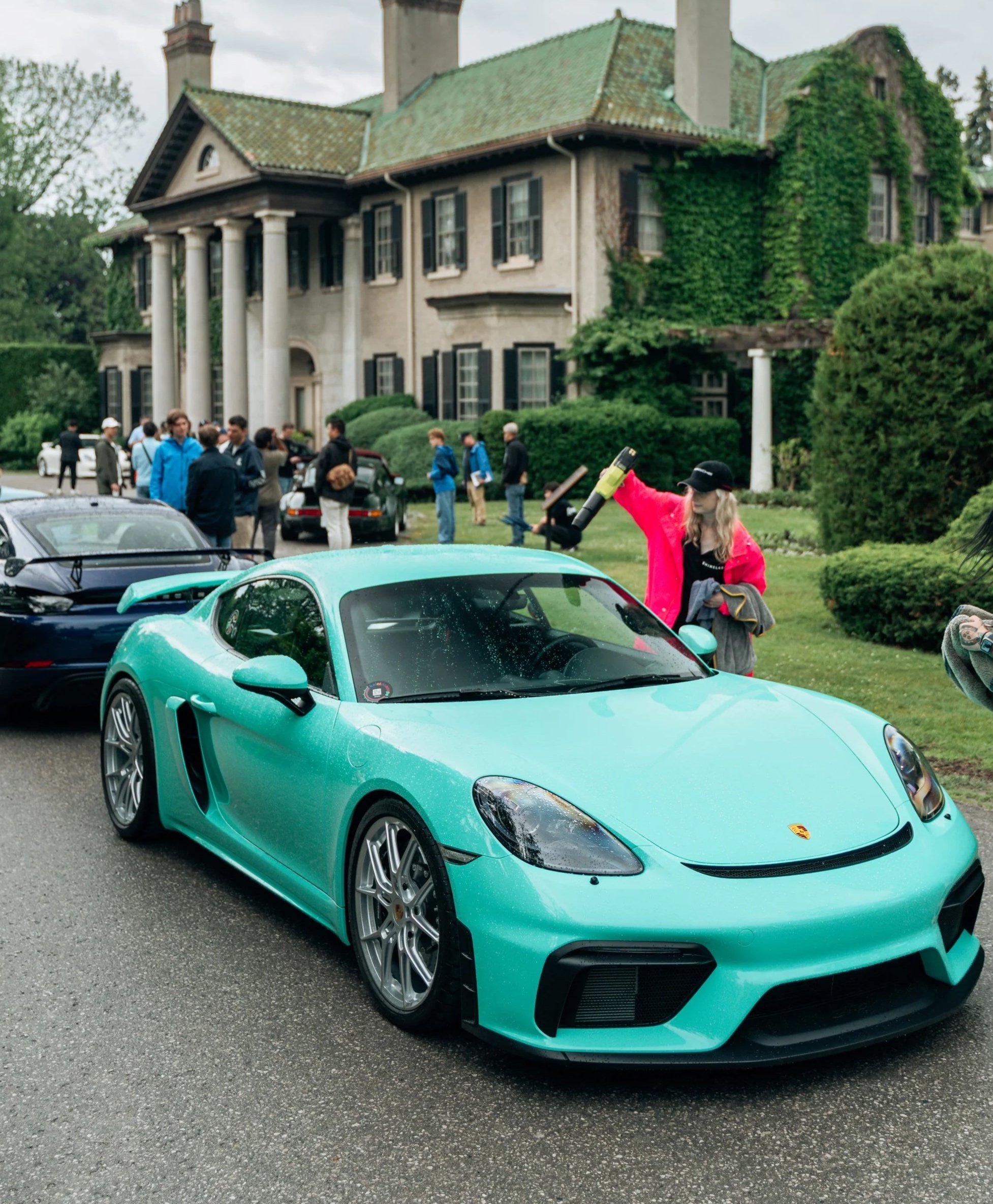A mint green sports car parked in front of a large old house with a green, moss-covered roof and ivy climbing on the side. Several people are gathered around the area. The car has a Porsche logo on the front.
