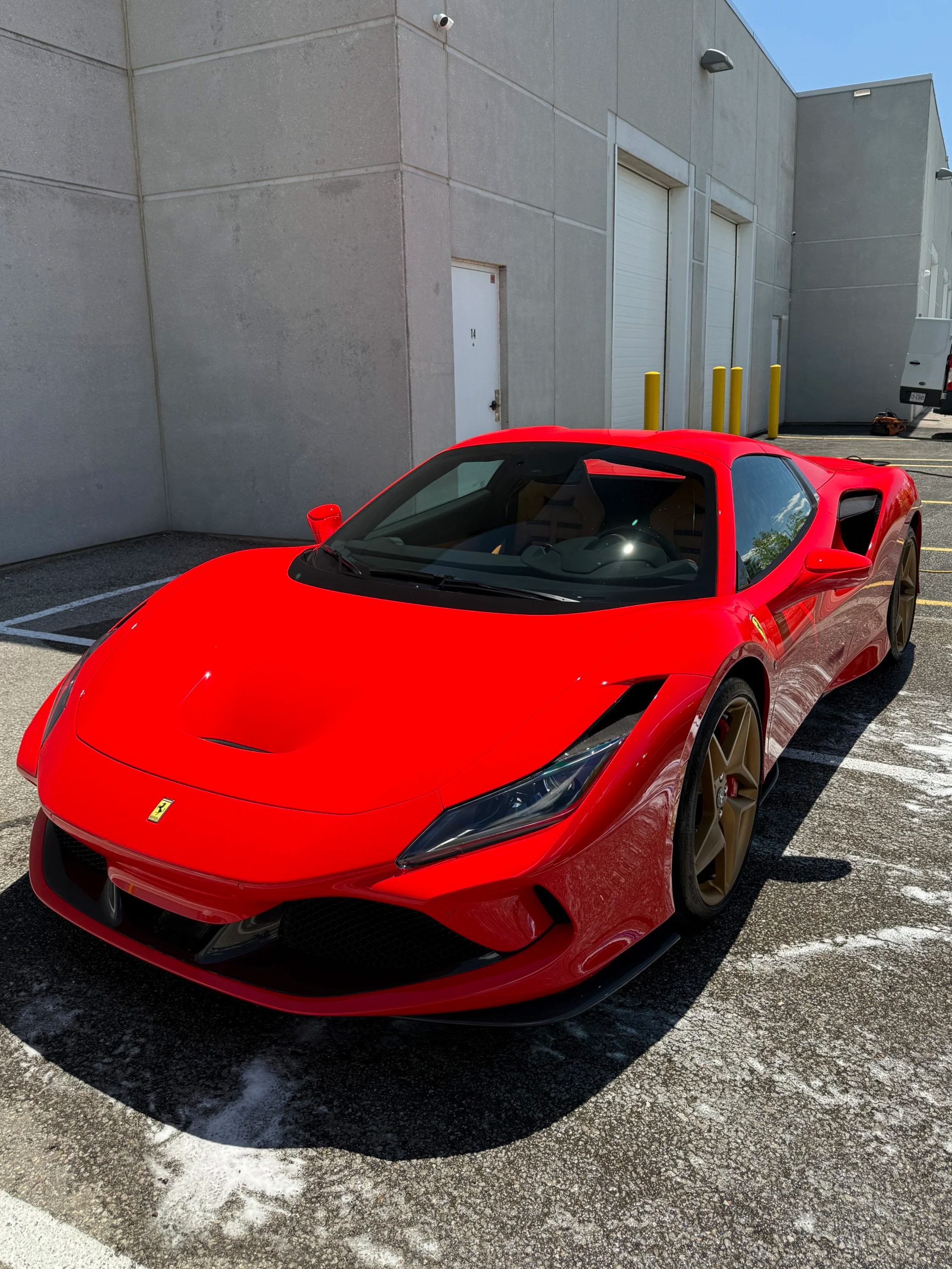 A red Ferrari sports car parked outside a gray industrial building with closed garage doors and yellow safety bollards.