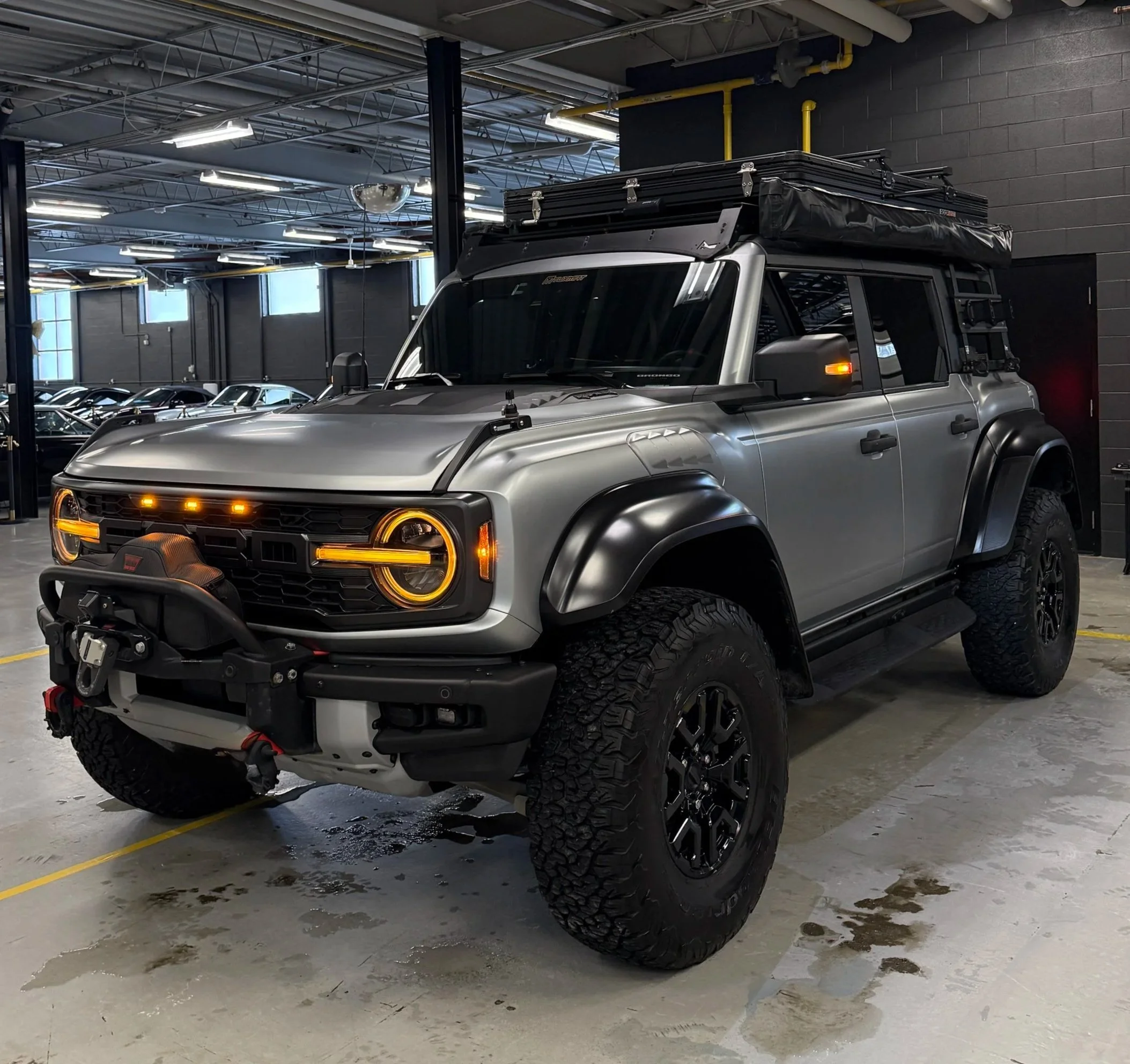 A silver off-road vehicle with yellow headlights and large tires parked inside a large industrial space with fluorescent lights and a high ceiling. The vehicle has a roof rack with a black cover and yellow pipes on the wall behind it.