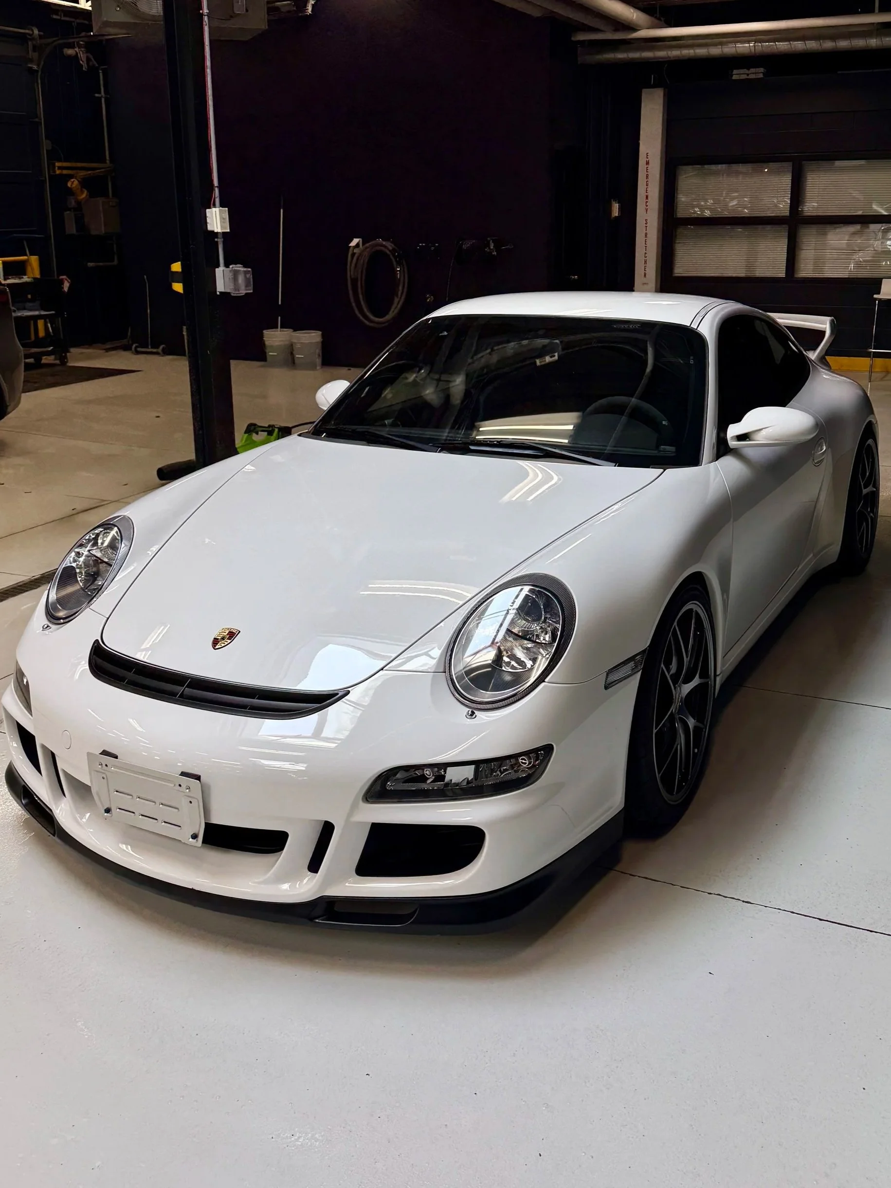 A white Porsche sports car parked inside a garage with a black interior and various tools and equipment on the walls.