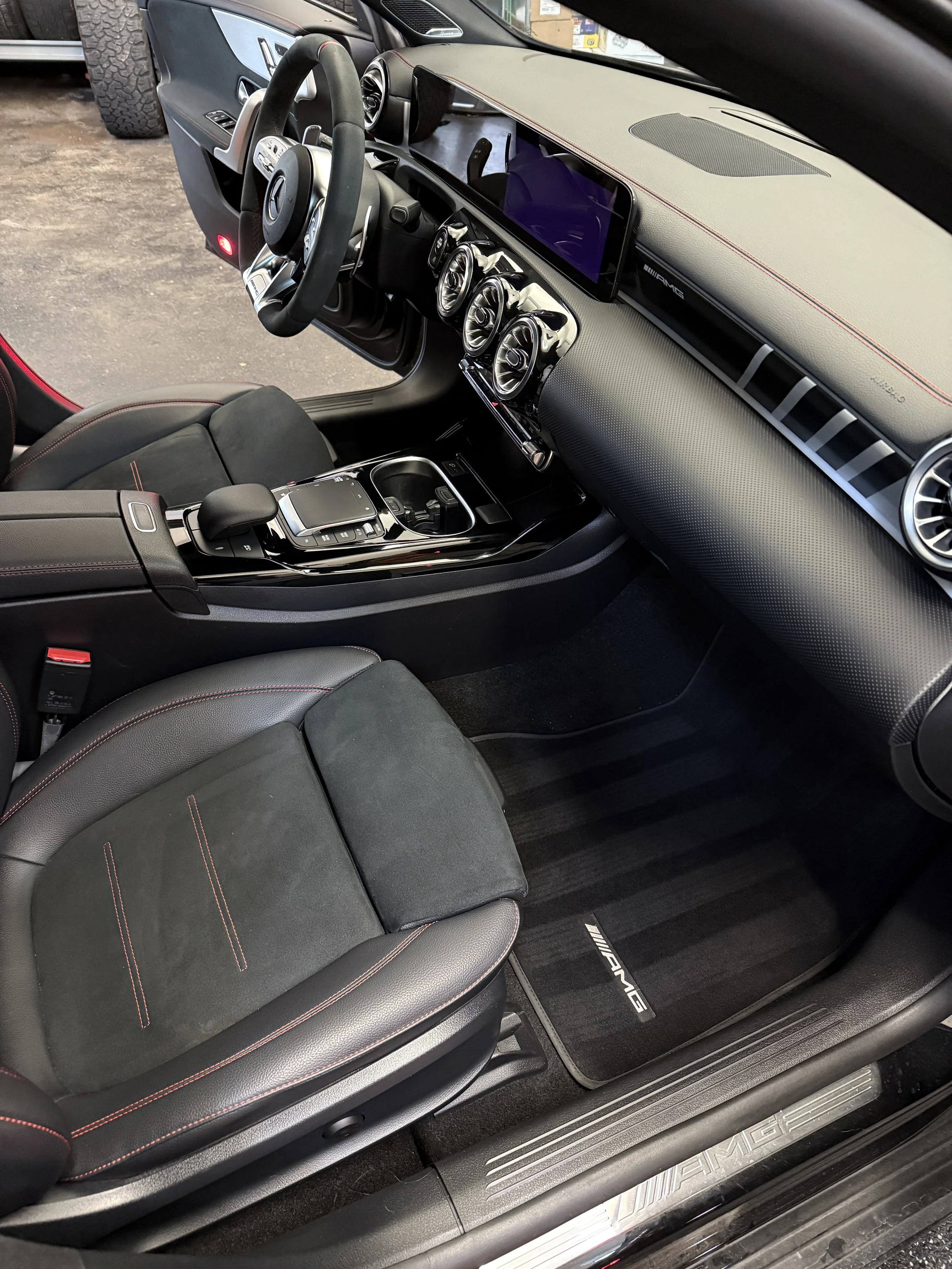 Interior of a luxury Mercedes-Benz vehicle, showing the driver's seat, steering wheel, dashboard, center console, and passenger seat, with AMG branding on the floor mat.