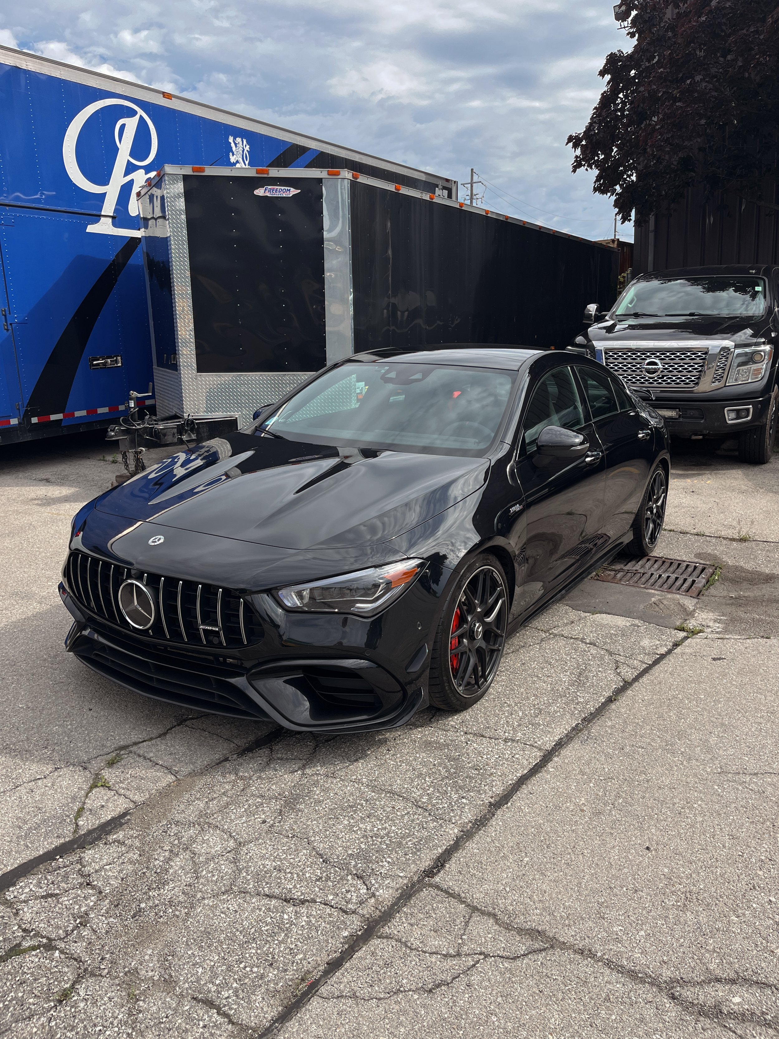 A black Mercedes-Benz car parked on a cracked asphalt lot, with a large blue trailer and a black pickup truck in the background. The sky is partly cloudy.