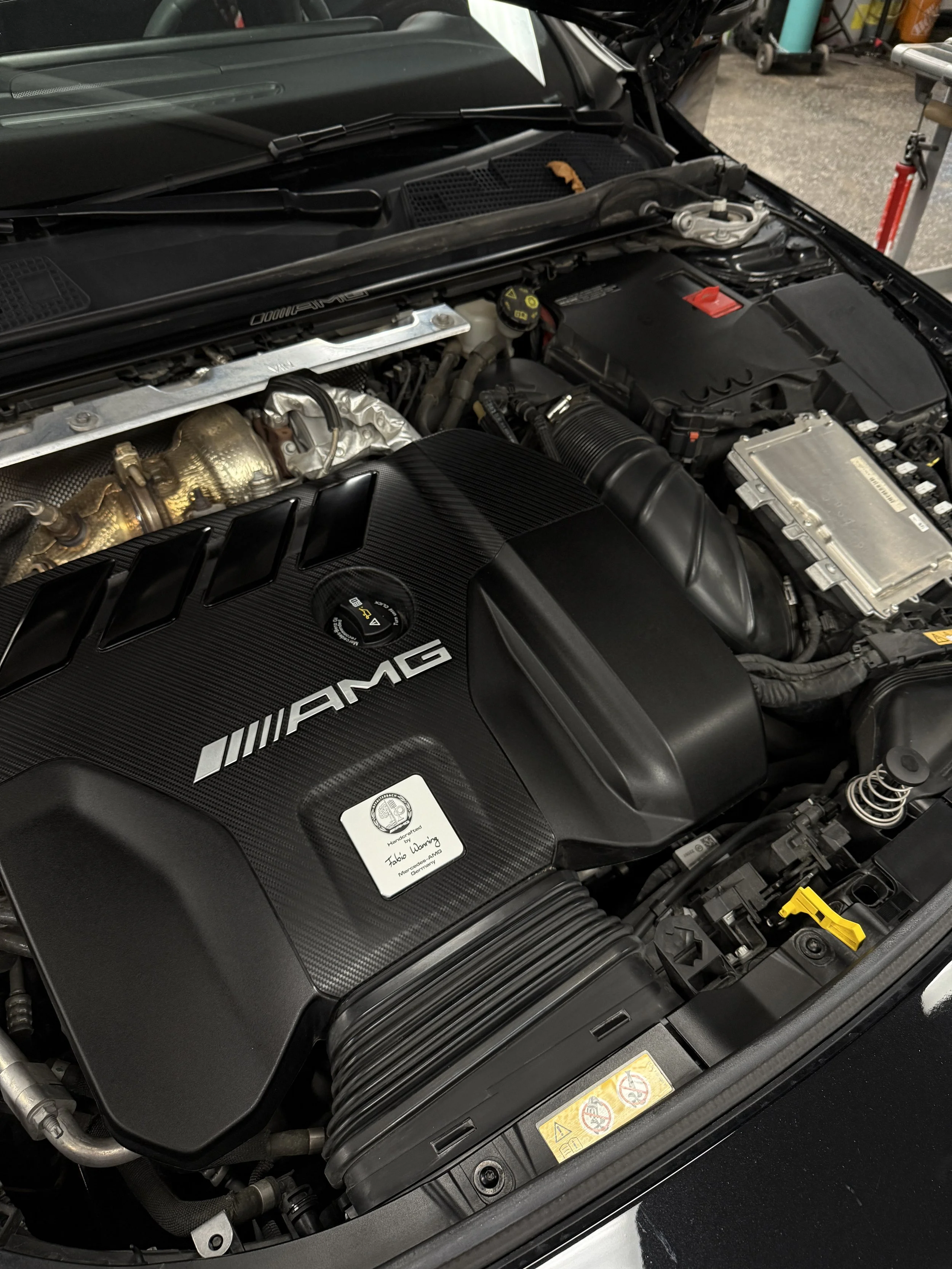 Close-up view of a Mercedes-AMG engine bay, featuring a carbon fiber engine cover with AMG logo, various components, and engine parts.