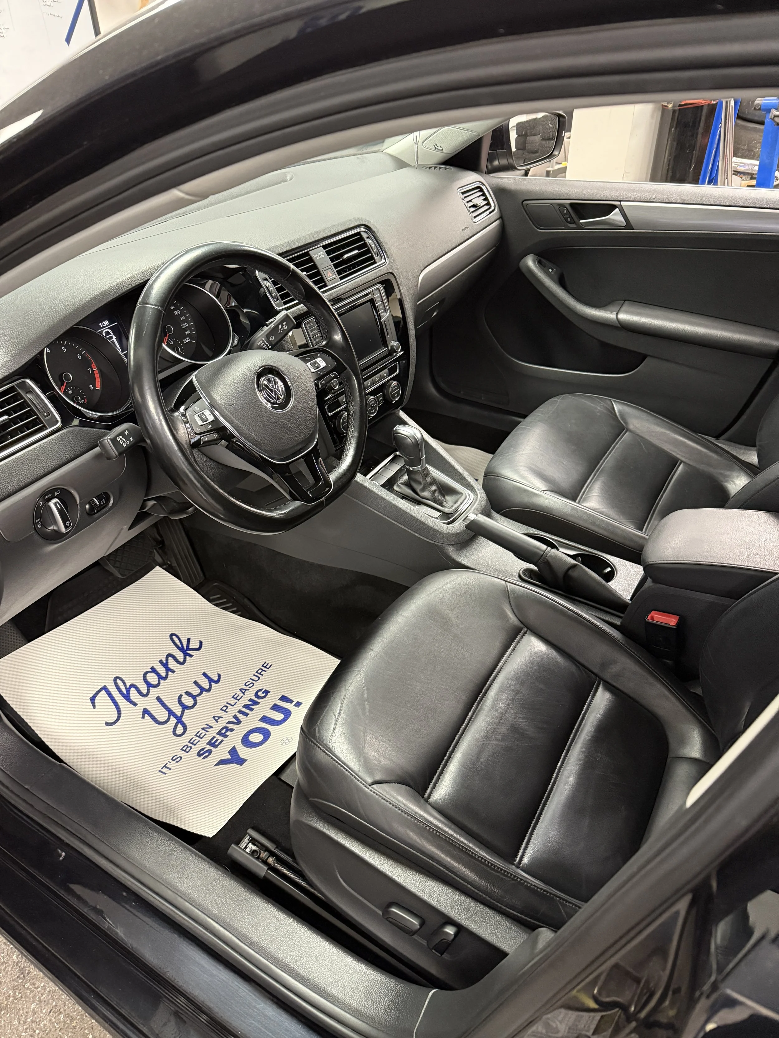Interior of a black Volkswagen car showing the front seats, dashboard, steering wheel, and center console with a paper mat on the driver's side that says, 'Thank you, it has been a pleasure serving you!'