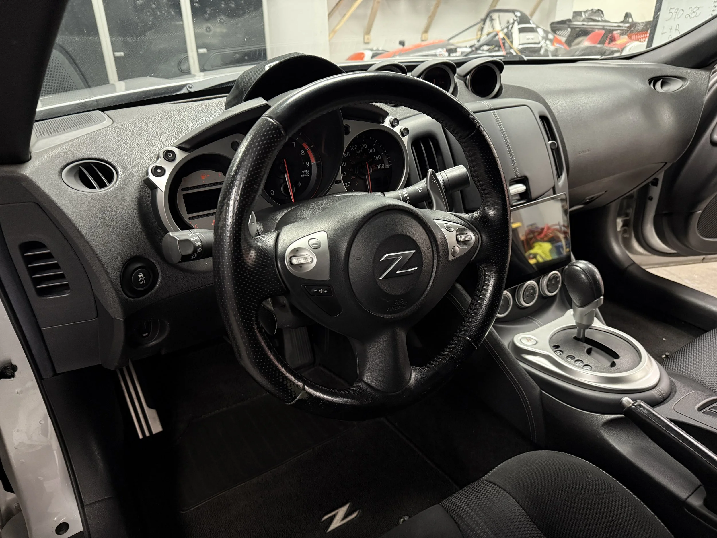Interior view of a Nissan 370Z, showing the black steering wheel, dashboard with gauges, and center console with gear shift and climate controls.