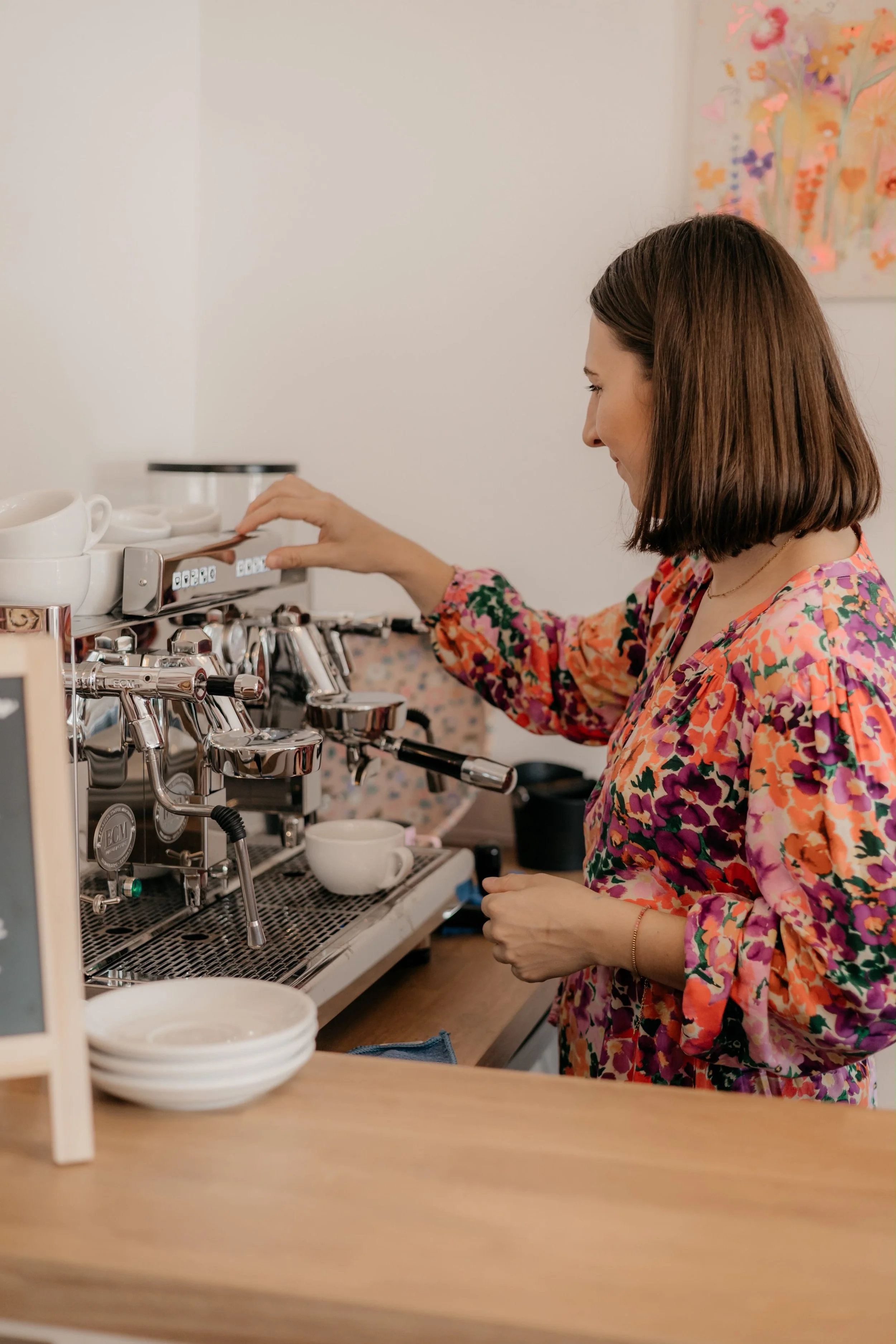 Frau bedient eine Espressomaschine in einem Café, trägt ein bunt gemustertes Kleid und hat braunes, schulterlanges Haar.