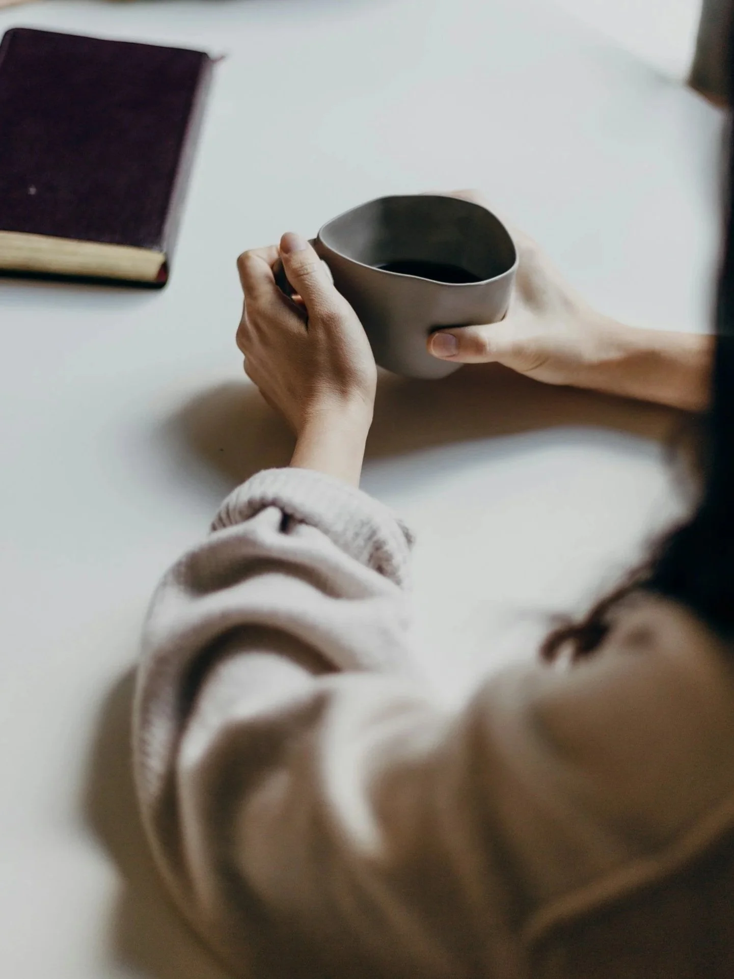 Person holding a gray ceramic mug with dark beverage, sitting at a white table, with a closed book nearby.