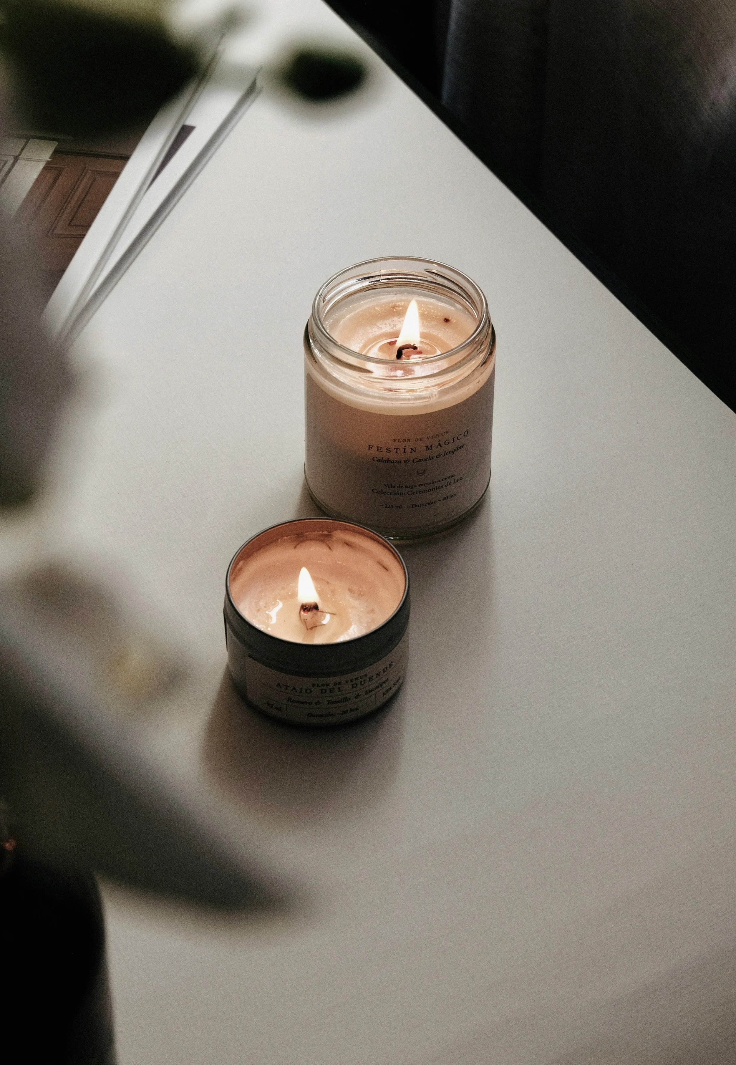 Two lit scented candles on a white surface, with a blurred dark object in the foreground and a portion of a kitchen in the background.