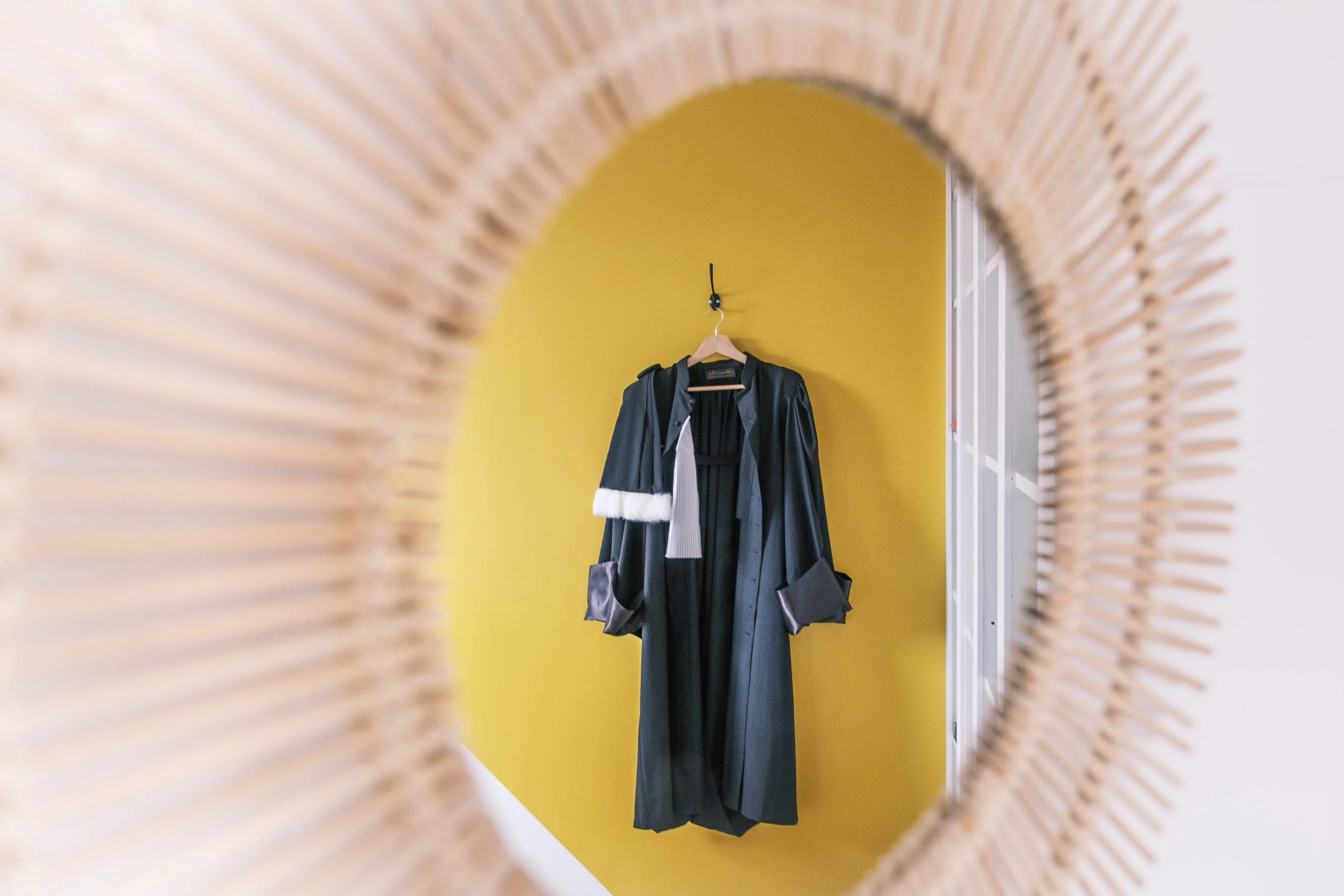Une robe longuement sombre, probablement noire, suspendue sur un porte-mantaux, vue à travers un cadre en rotin ou en bois tressé, avec un mur jaune en arrière-plan.