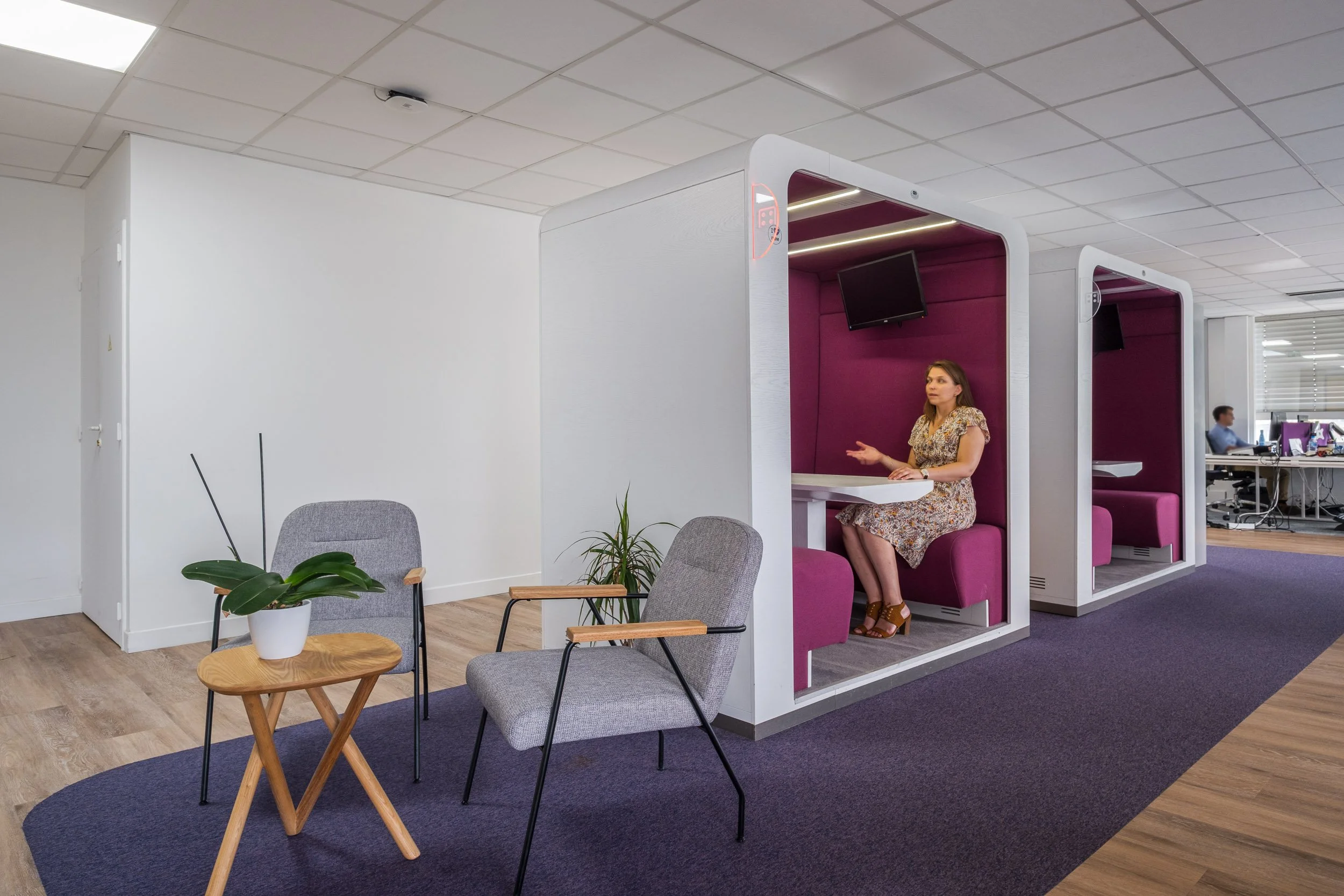 Espace de travail avec des cabines individuelles de style moderne, couleurs blanches et accents roses, une femme assise à l'intérieur d'une cabane et un homme travaillant à un bureau en arrière-plan, environnement de bureau contemporain.