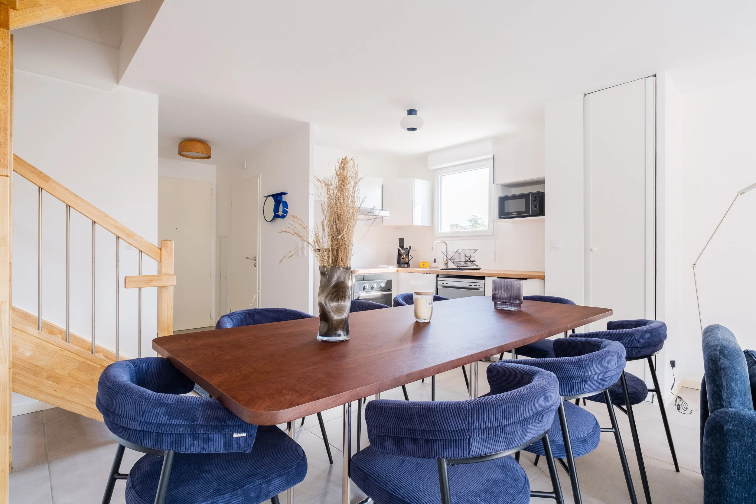 Salle à manger avec un grand tableau en bois, des chaises bleues en tissu, un vase avec des plantes séchées, et une cuisine en arrière-plan avec une fenêtre lumineuse.