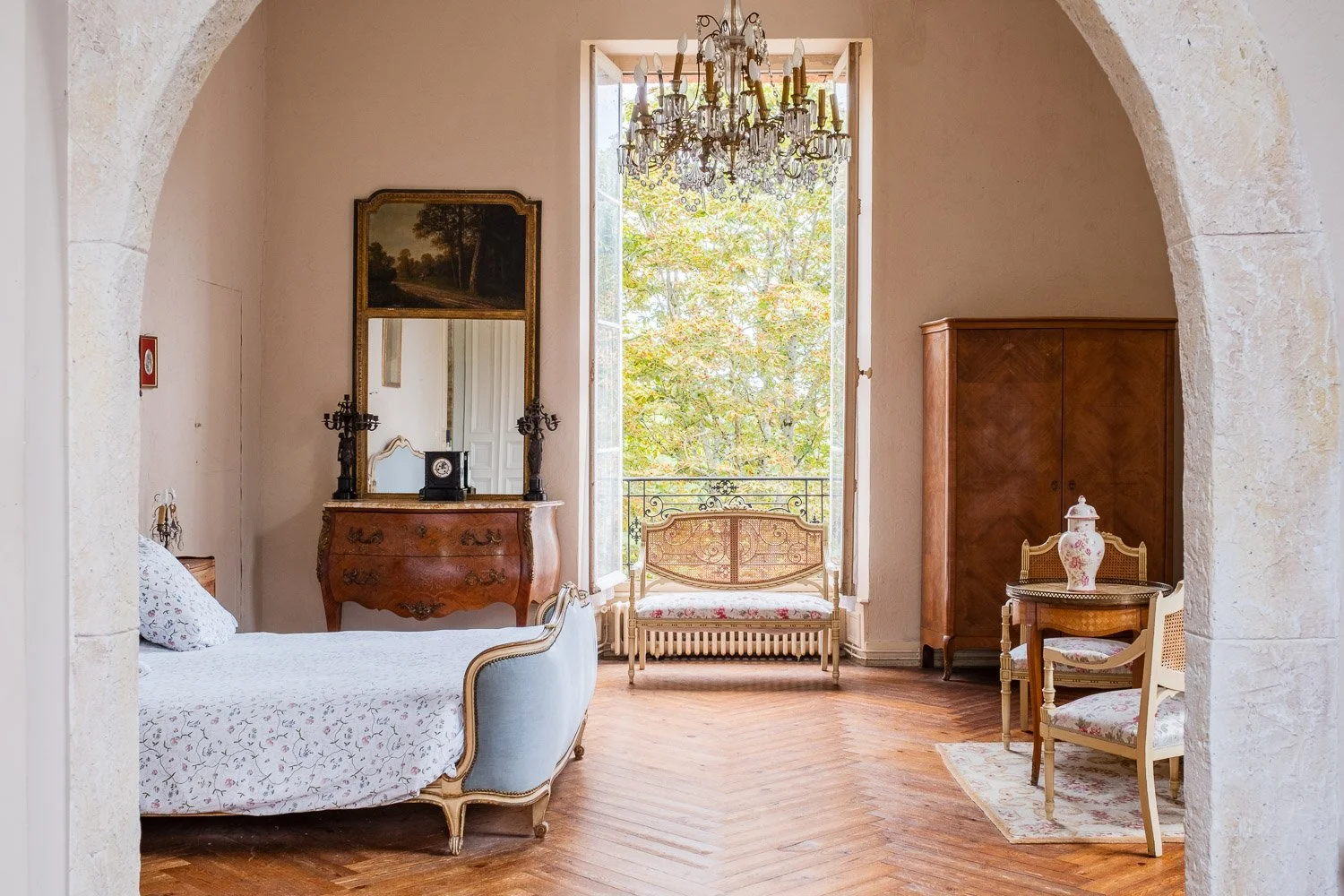 Salon avec mobilier ancien, canapé, armoire en bois, lampe, miroir, chandelier suspendu, fenêtre avec vue sur des arbres