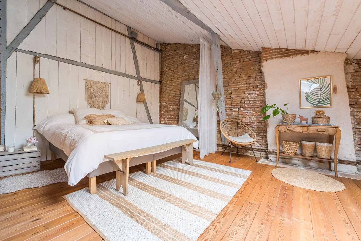 Chambre à coucher avec lit en bois, décorée de style rustique, avec mur en briques, tapis rayé et meubles en bois naturel, plantes et tableaux végétaux.