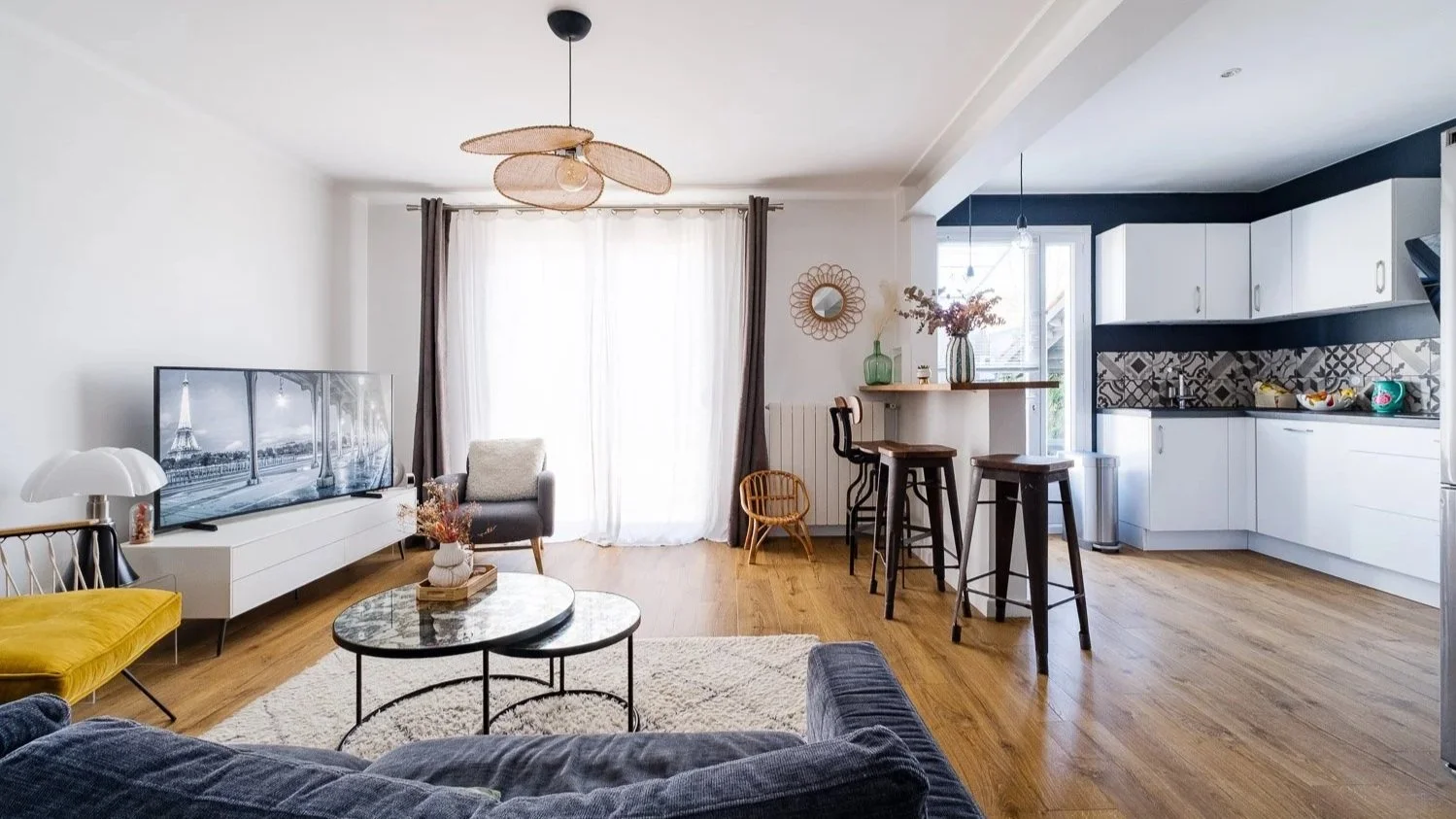 Intérieur d'un salon et cuisine modernes avec mobilier en bois, canapé gris, fauteuil beige, TV, espace repas avec tabourets en bois et cuisine blanche avec comptoir en noir, décorations florales et paniers en osier.