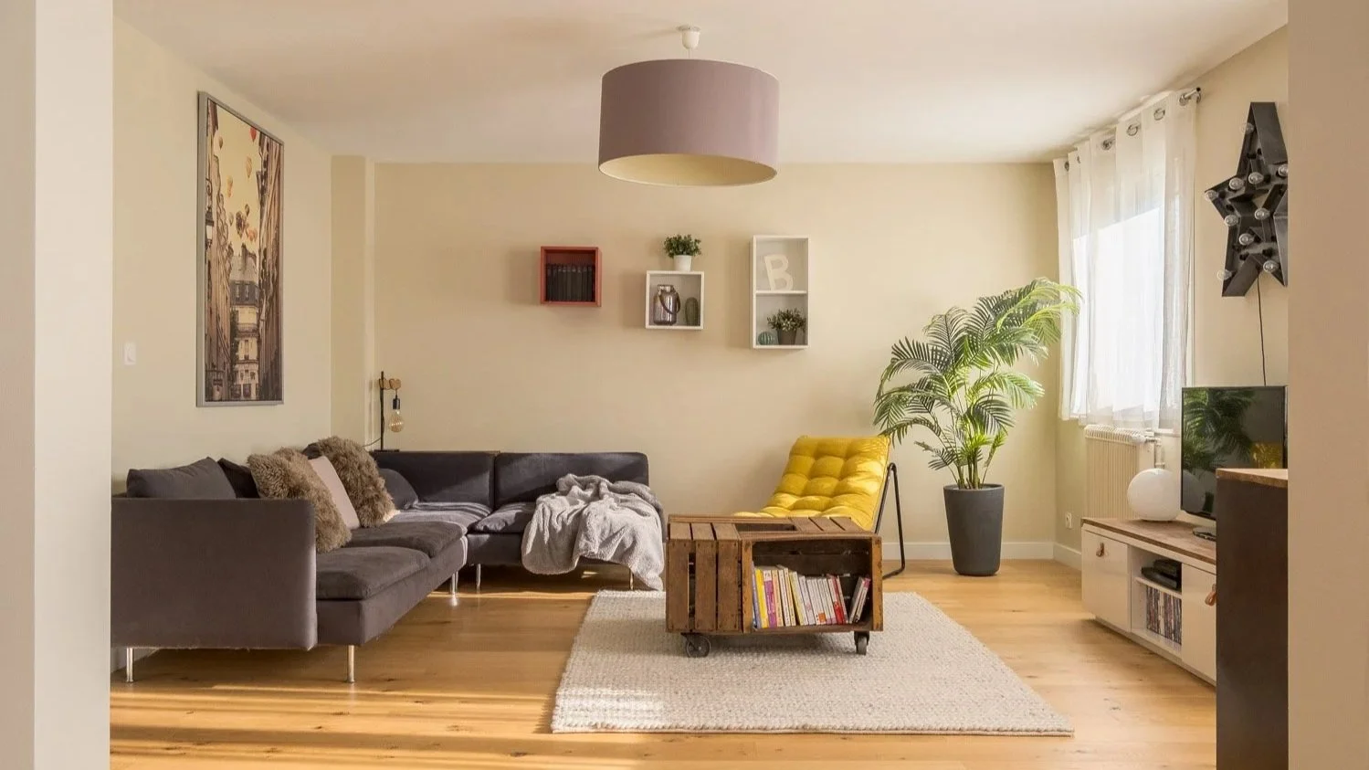 Salon moderne avec canapés gris, table en bois, fauteuil jaune, télévision, plantes vertes, étagères murales, lampes et rideaux blancs.