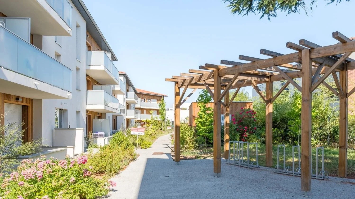 Résidence moderne avec balcons en façade, espace extérieur avec pergola en bois, plantes et fleurs, chemin piétonnier.