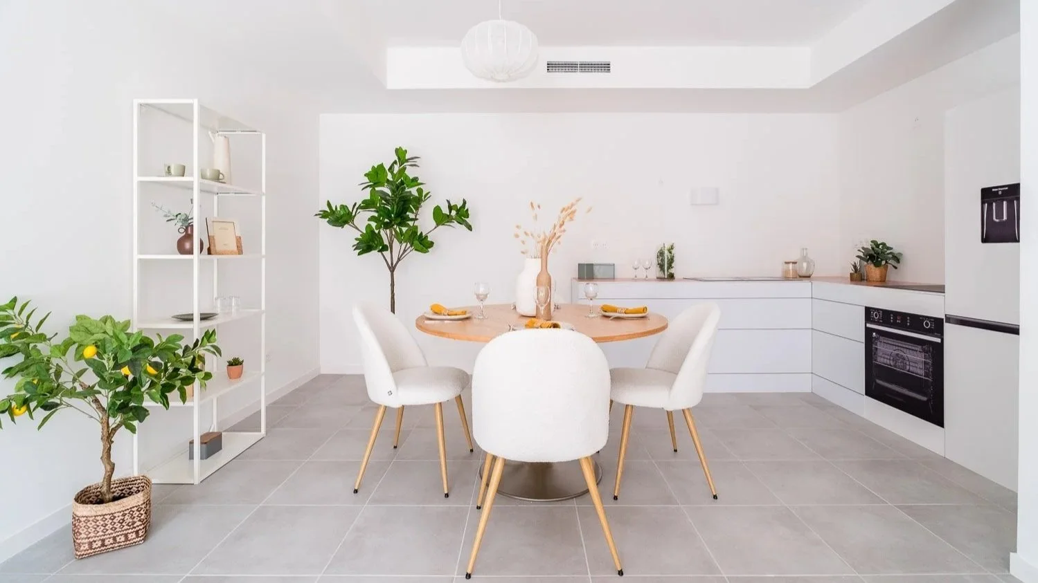 Cuisine moderne avec table ronde, chaises blanches, étagère blanche, plantes vertes, et appareils électroménagers encastrés en blanc.